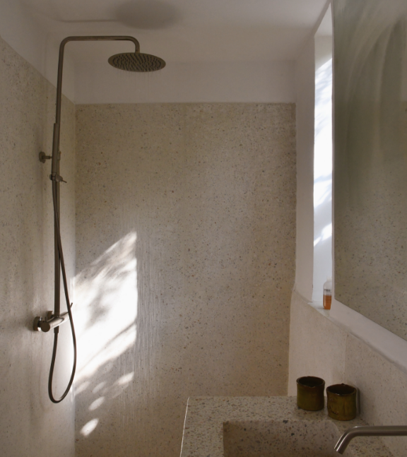 Douche avec mur en béton, pommeau de douche rond, et deux petits pots en terre cuite sur une surface en béton à côté d'une fenêtre.