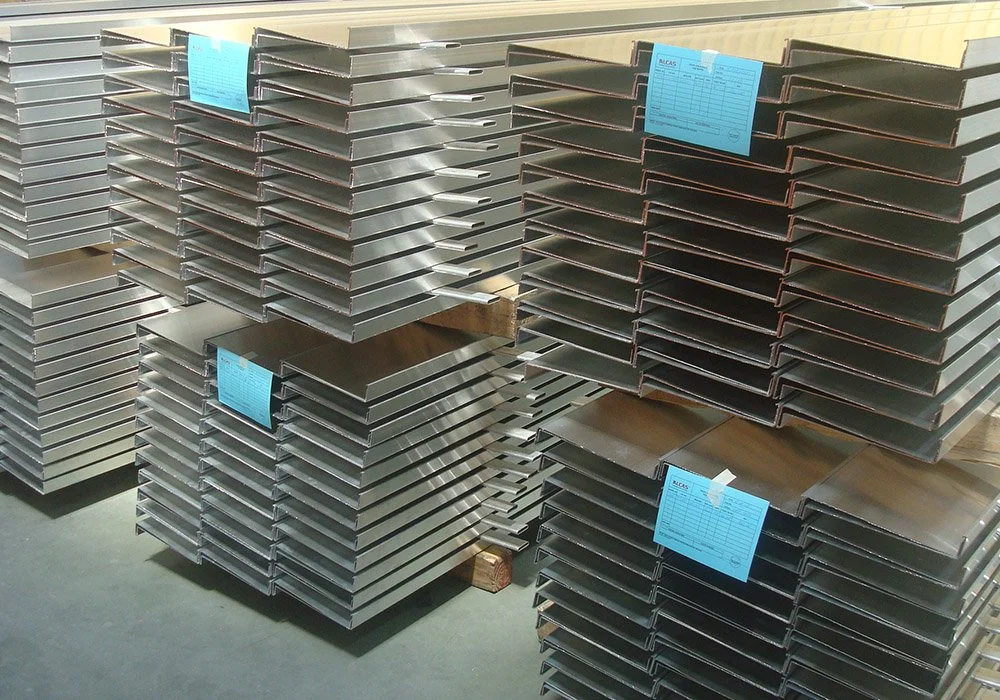Stacks of metal channels or brackets with blue labels on each stack, arranged on wooden pallets in an industrial setting.