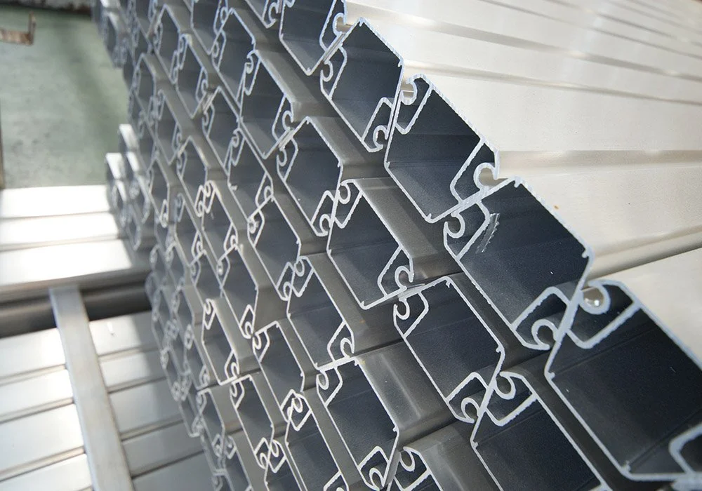 Stacked metal extrusion profiles with rectangular hollow sections and intricate edge details, arranged on a warehouse shelf.