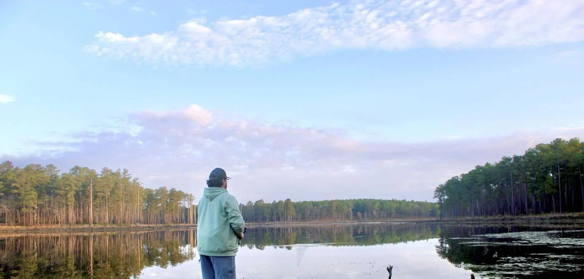 A person standing by a calm lake, surrounded by trees, under a partly cloudy sky.