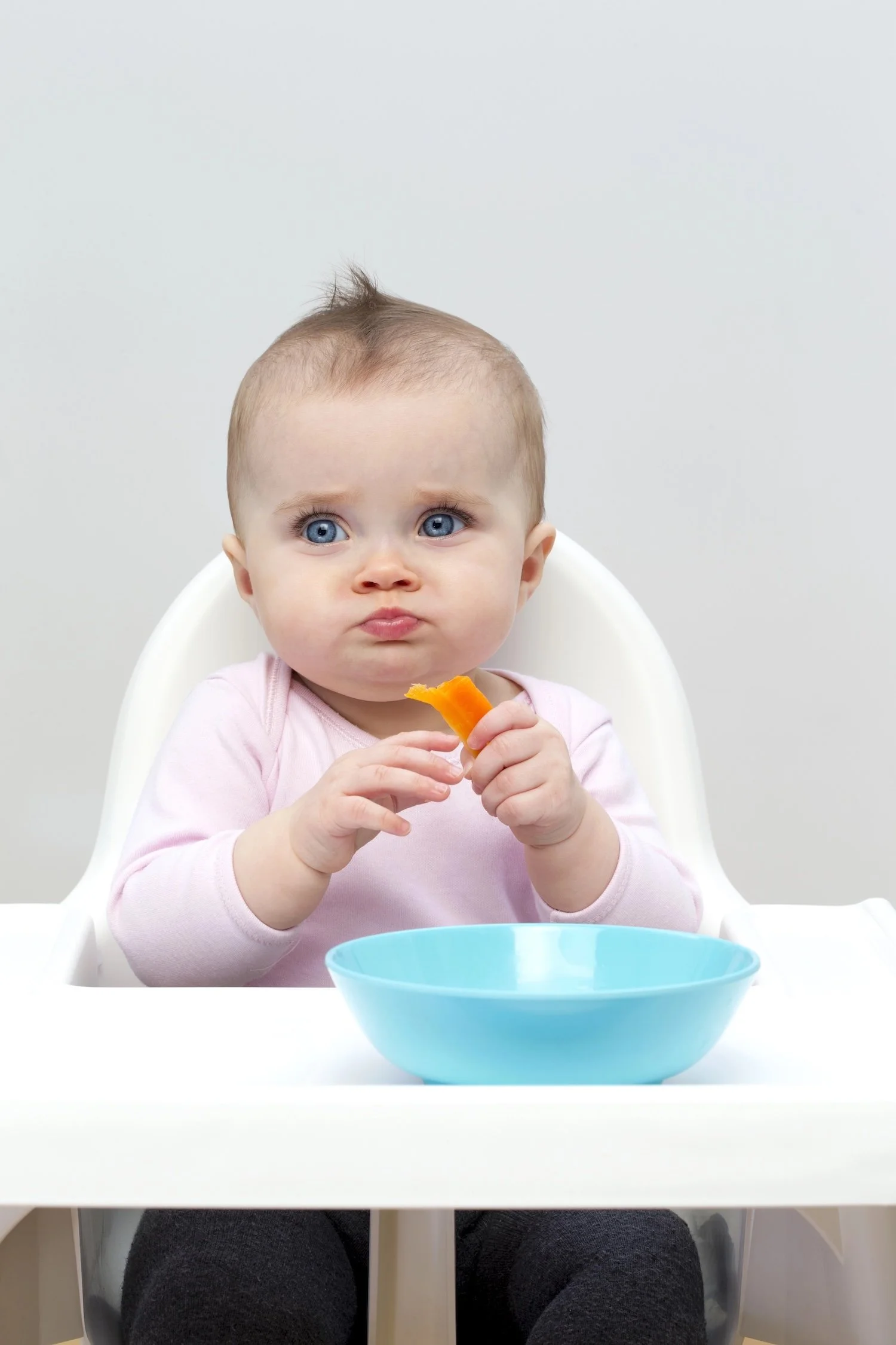 A young child with blue eyes and light brown hair in a high chair at a white table, holding an orange slice, with a blue bowl in front.