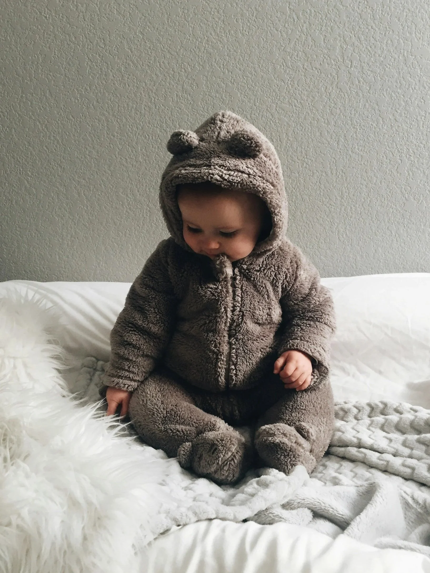 A young child dressed in a fuzzy bear costume, sitting on a white bed with a textured beige wall in the background.