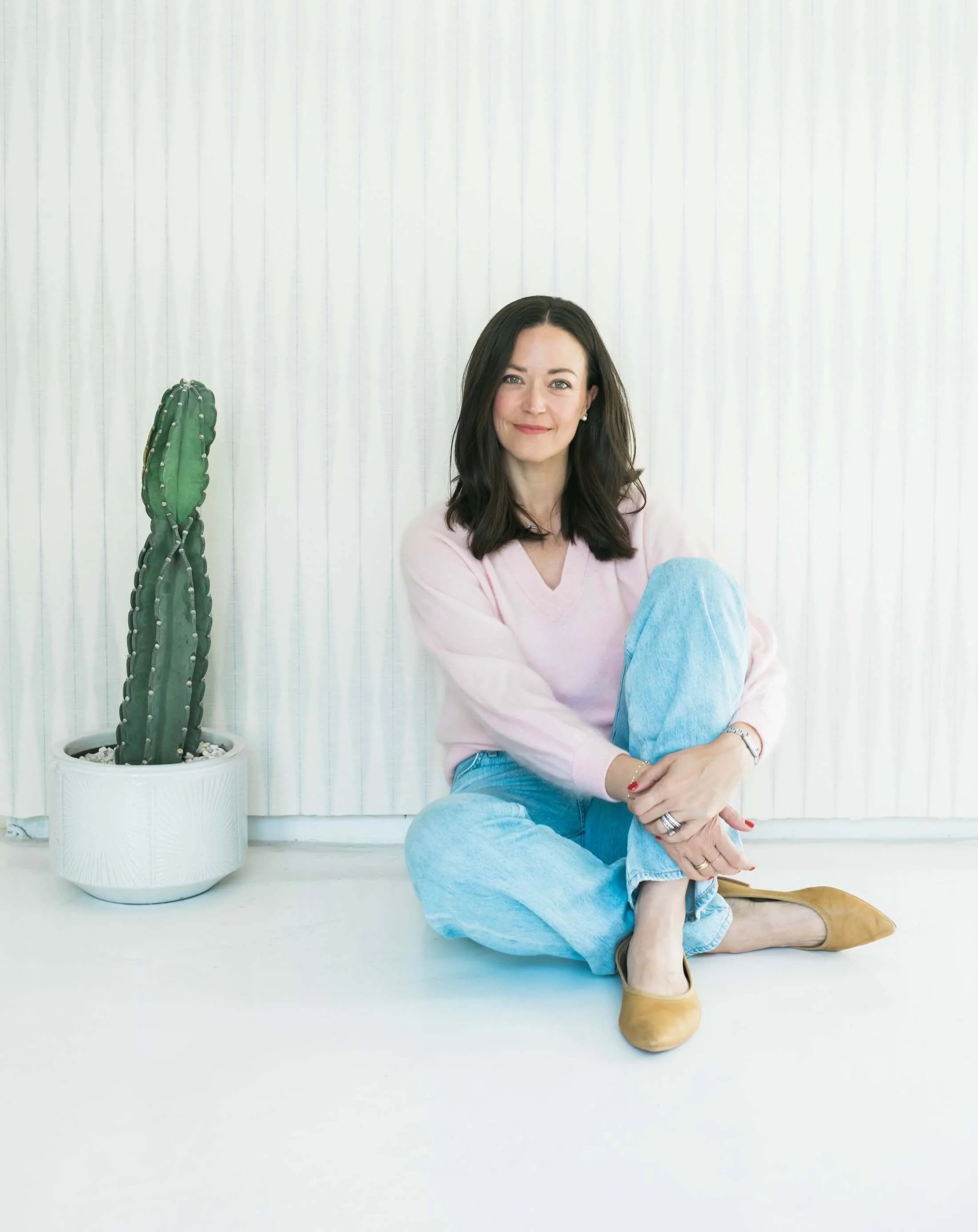 Amy Onderdonk is sitting on the floor with her legs crossed, dressed in a light pink sweater, blue jeans, and tan boots, smiling at the camera. There is a tall green cactus in a white pot to her left and a white vertical striped background.