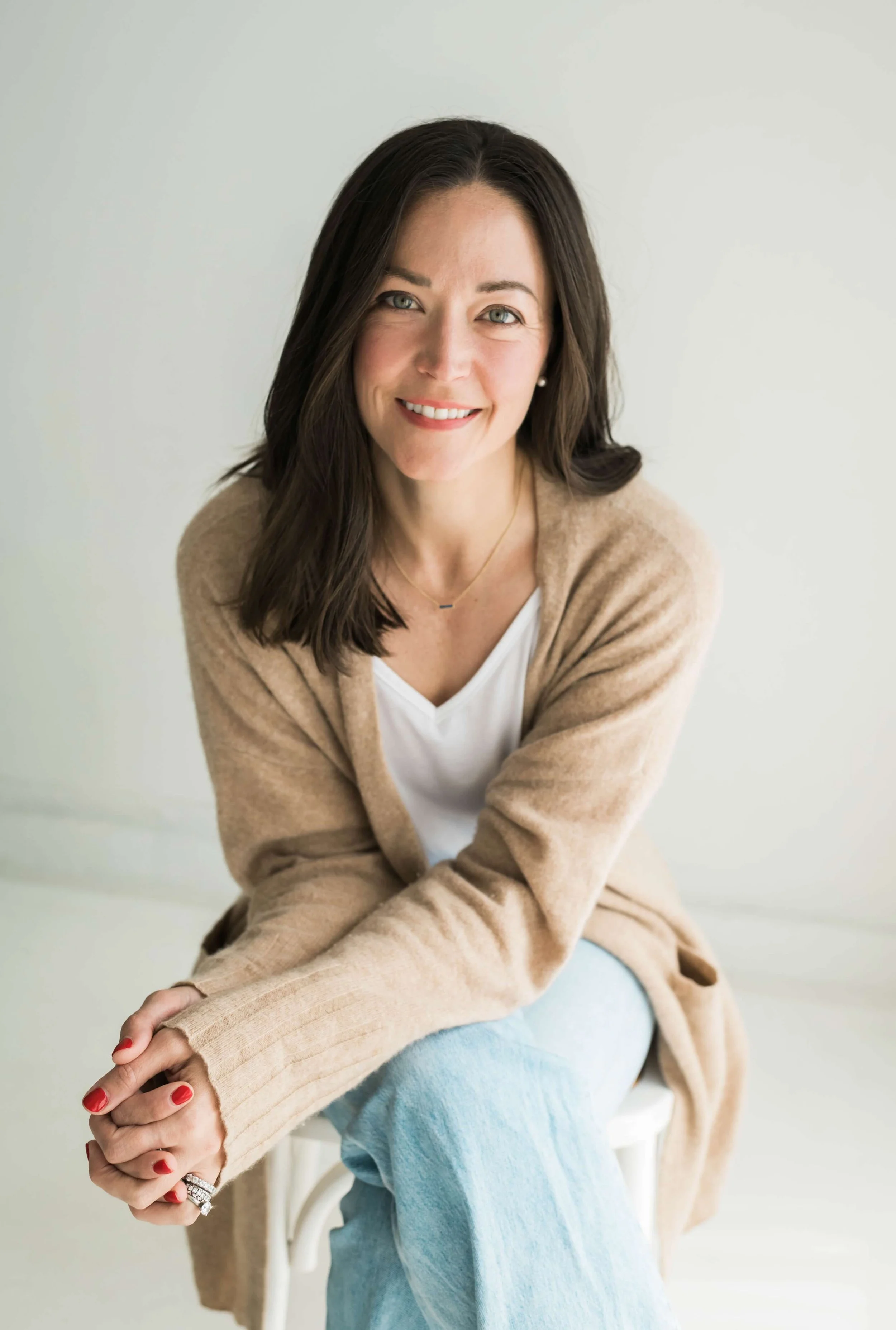 Amy Onderdonk, with a warm smile, is sitting on a white chair against a plain light background. She is wearing a beige cardigan over a white top, light blue jeans.