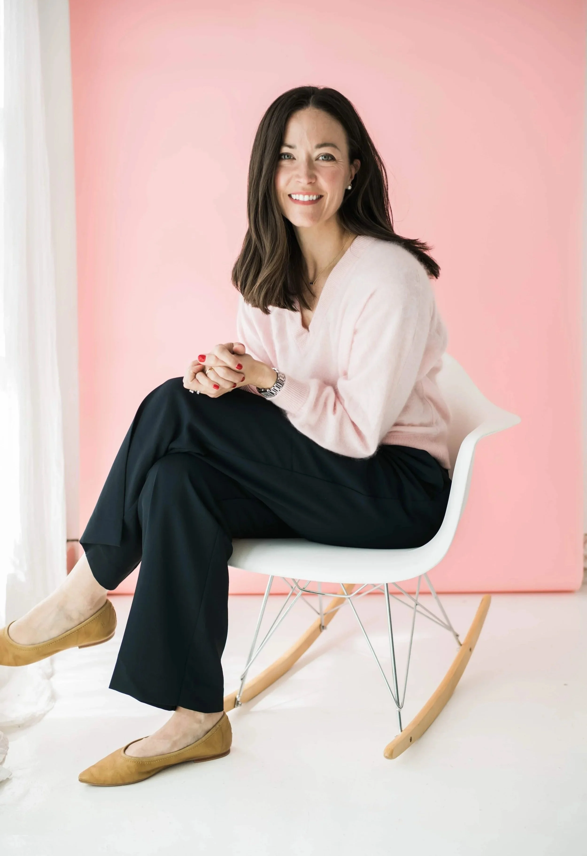Amy Onderdonk is sitting on a white chair within a room with a pink wall background. She is wearing a light pink sweater, black pants, and tan flats. She has dark brown hair, is smiling, and has a watch on her left wrist.