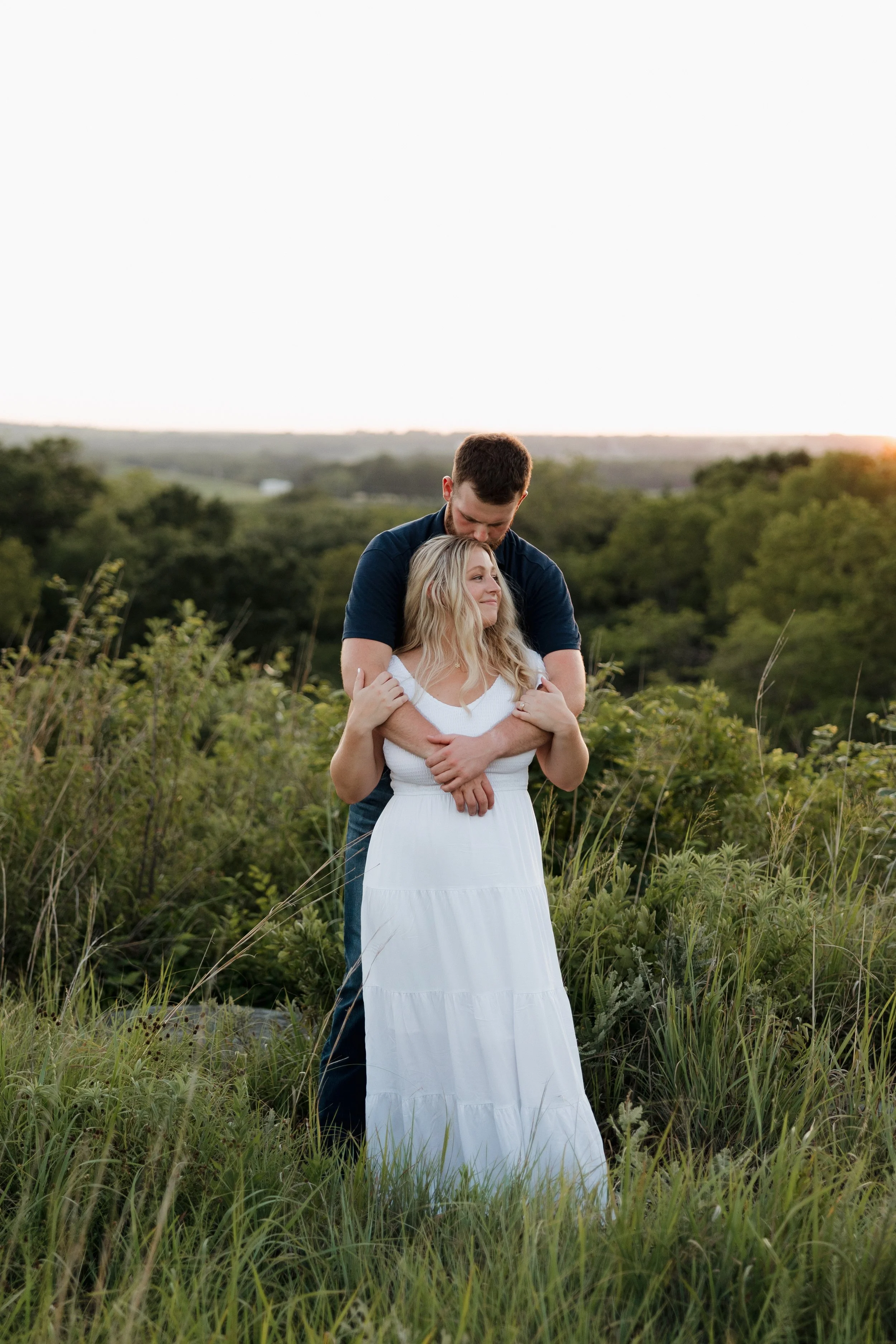 A couple embracing in a grassy field during sunset, with trees and hills in the background.