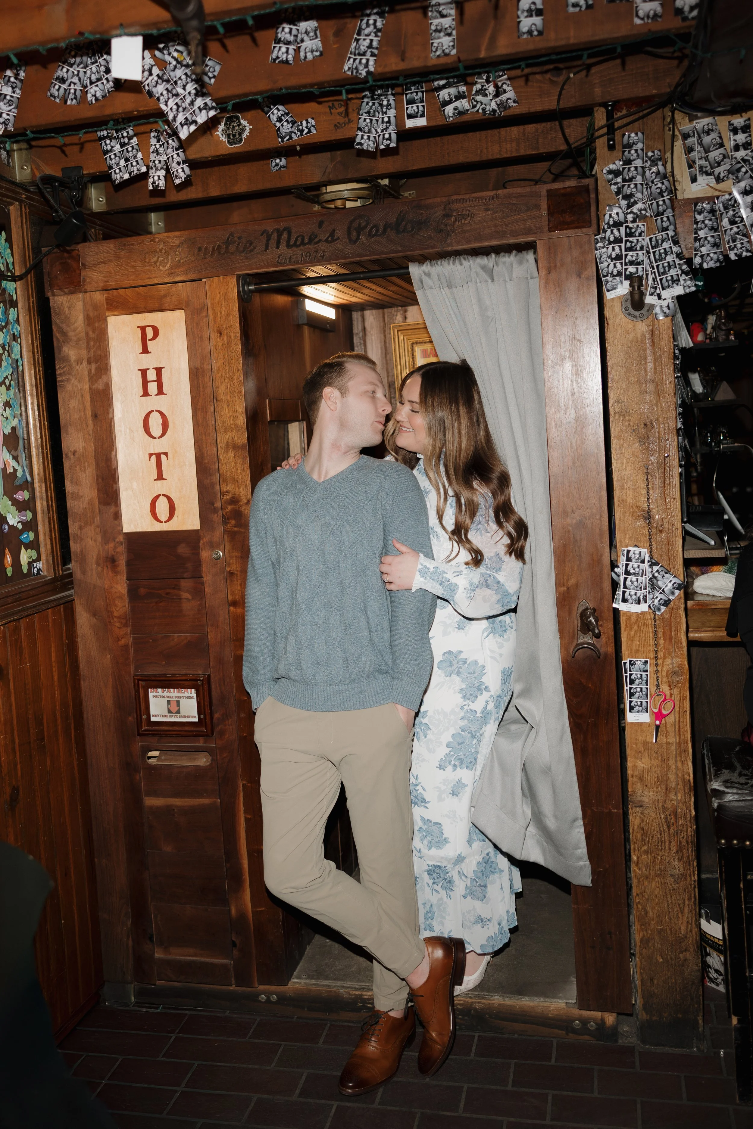A man and woman are posing in a photo booth, standing close with their faces touching and smiling. The man has short blonde hair and is wearing a gray sweater, tan pants, and brown shoes. The woman has long, wavy brown hair and is wearing a white dress with blue floral patterns and white shoes. The photo booth has a wooden frame with a