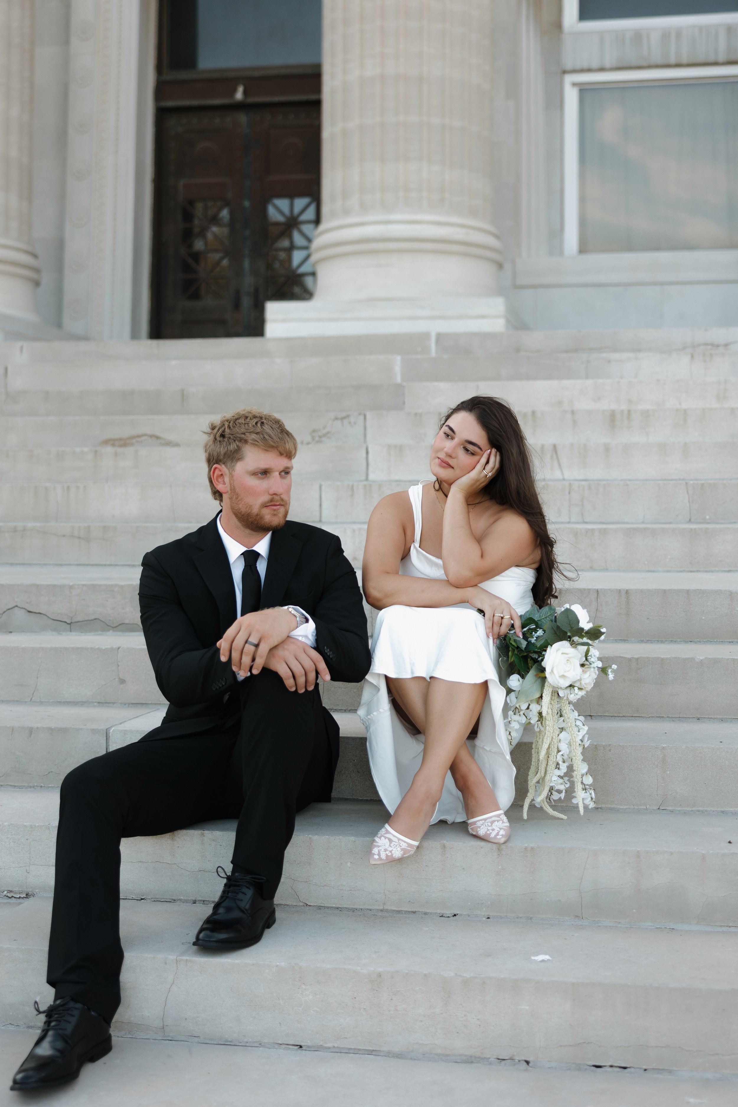 A bride and groom sitting on the steps outside a large building, with the groom wearing a black suit and the bride in a white dress, holding a bouquet of white roses.