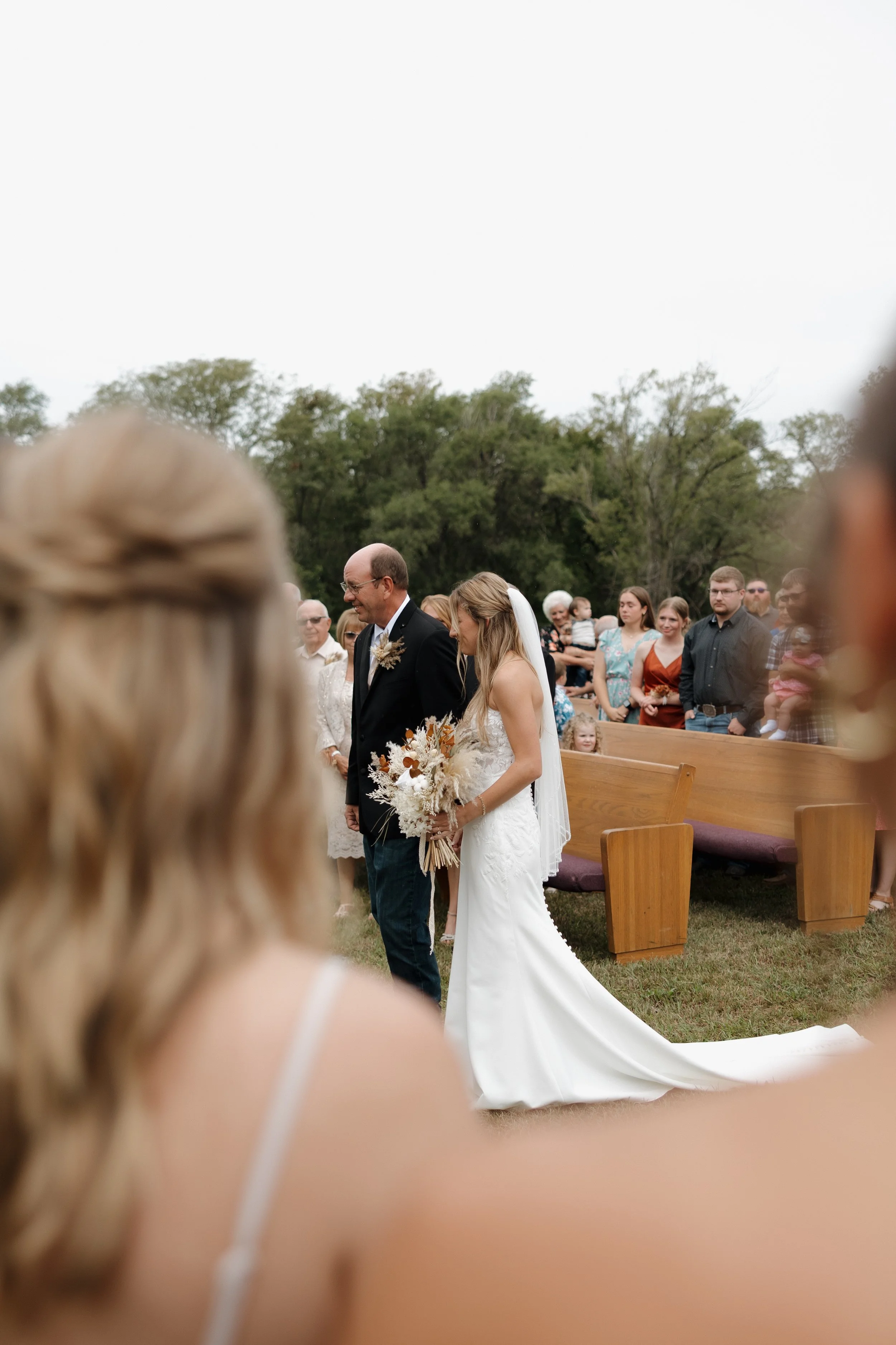 A bride in a white wedding dress and veil walking down the aisle looking at a man, possibly her father, at an outdoor wedding ceremony with guests in the background. The scene is set on a grassy area with trees and outdoor benches.