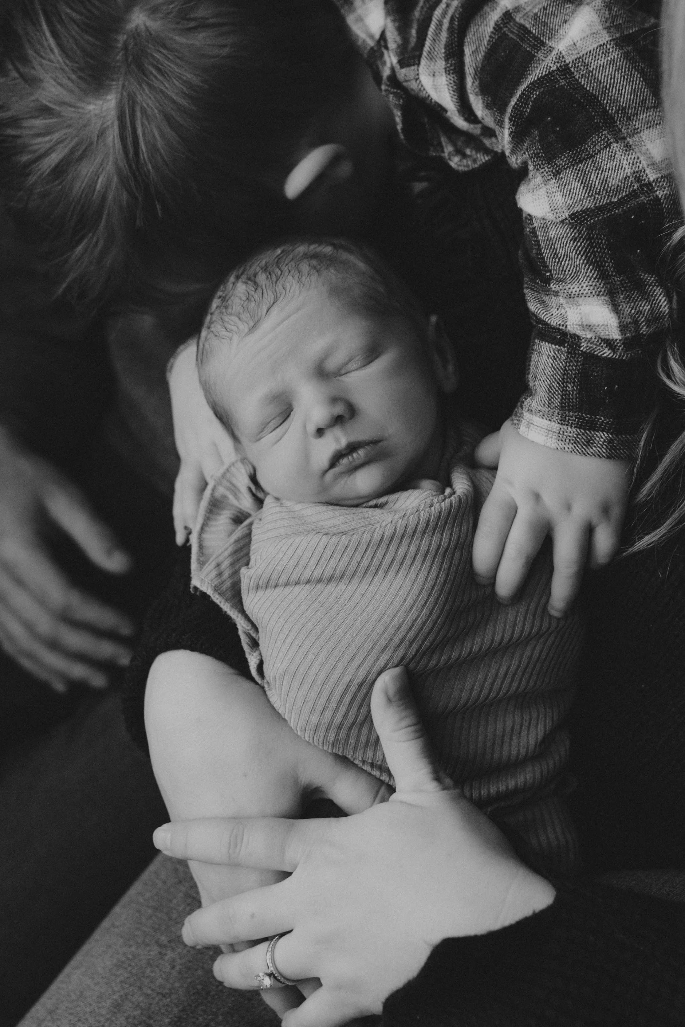A woman is gently holding a sleeping baby close to her chest, cradling the baby's head with her hand. The baby has closed eyes and a peaceful expression. The woman wears a ring on her finger and a plaid shirt, and there are other hands and arms around them.