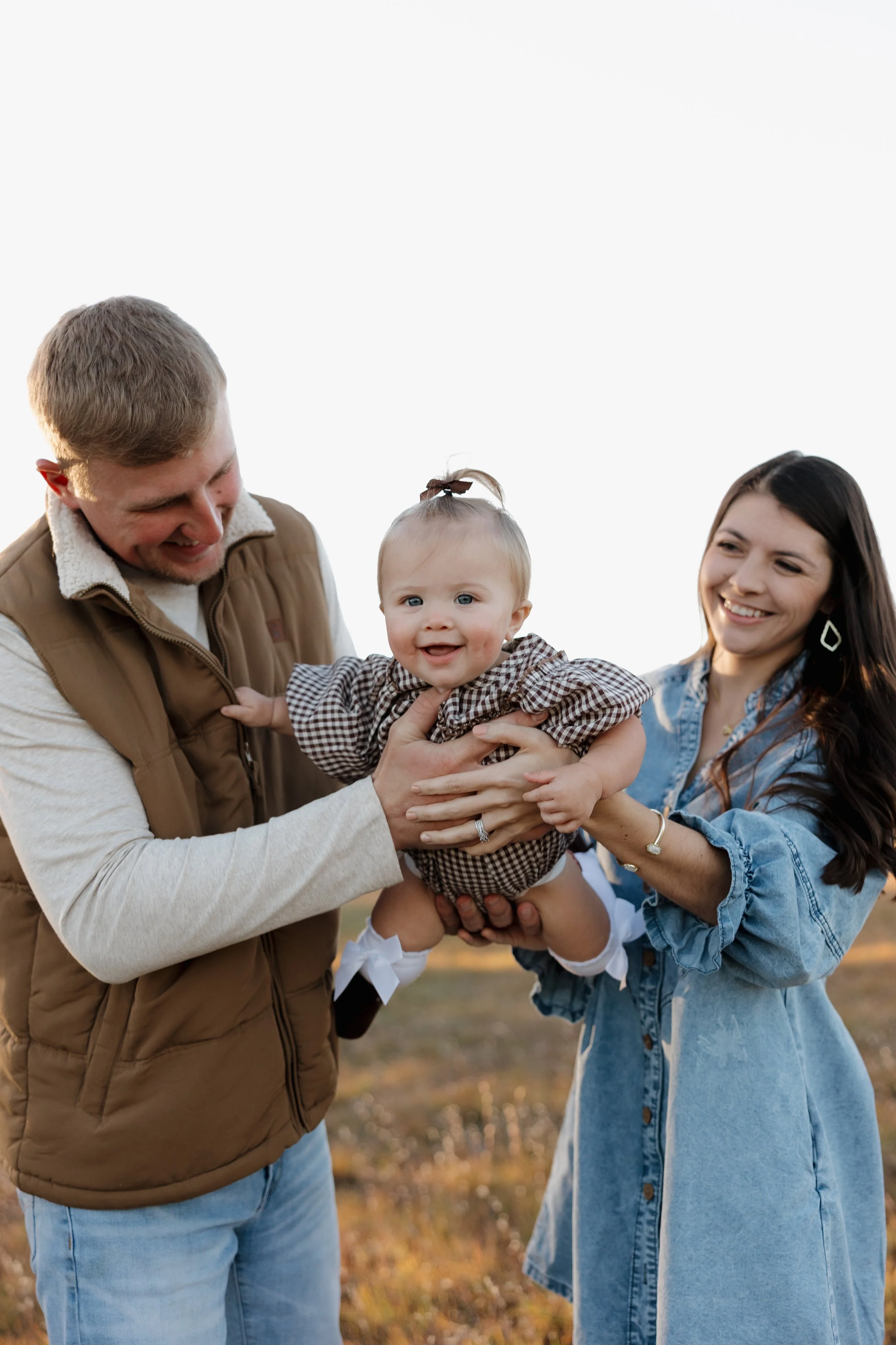 A happy family of three holding a smiling baby girl outdoors during sunset, with trees blurred in the background.