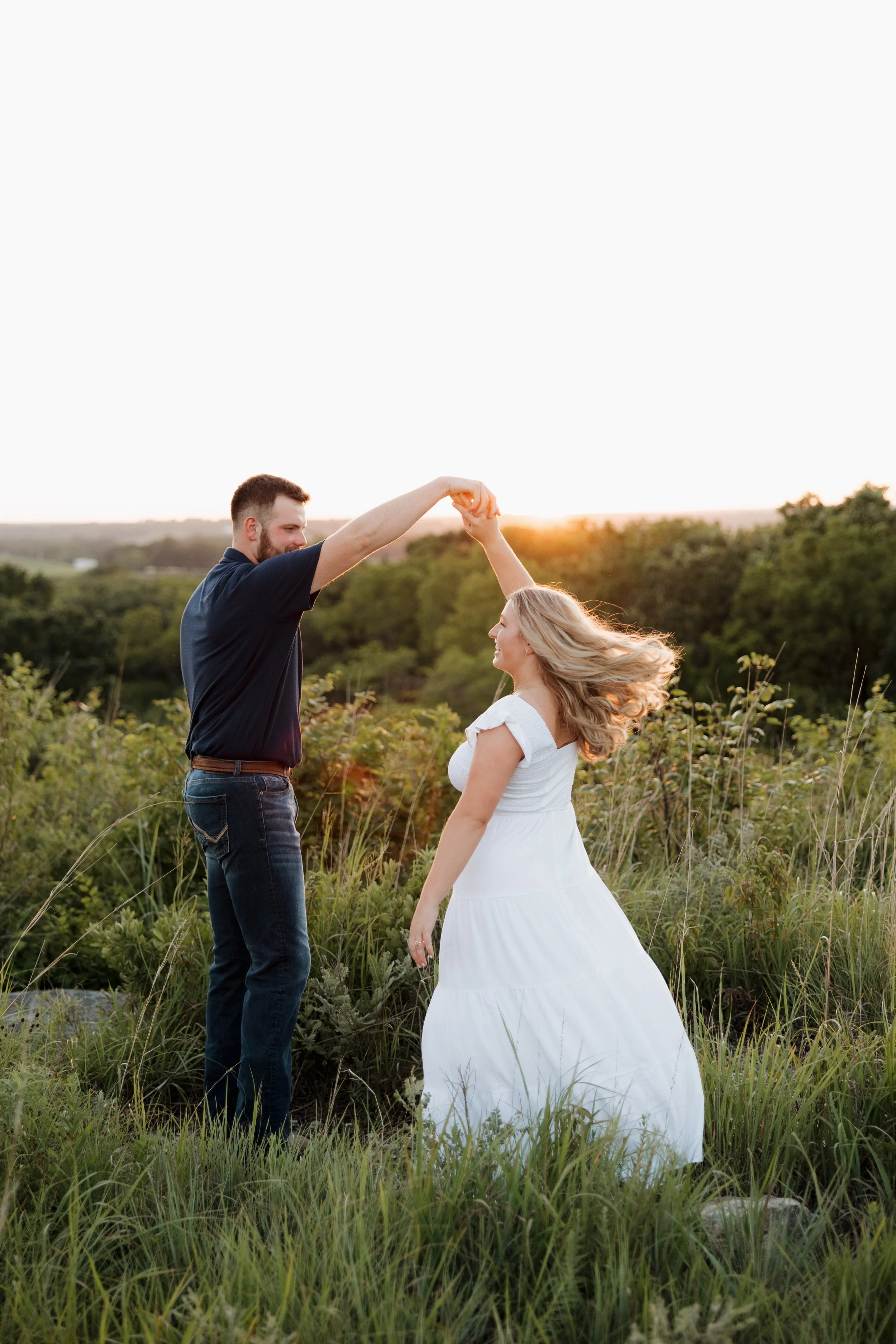 A couple dancing outdoors during sunset; the man is holding the woman’s hand as they twirl, with the woman in a white dress and the man in dark clothing, surrounded by green grass and trees.