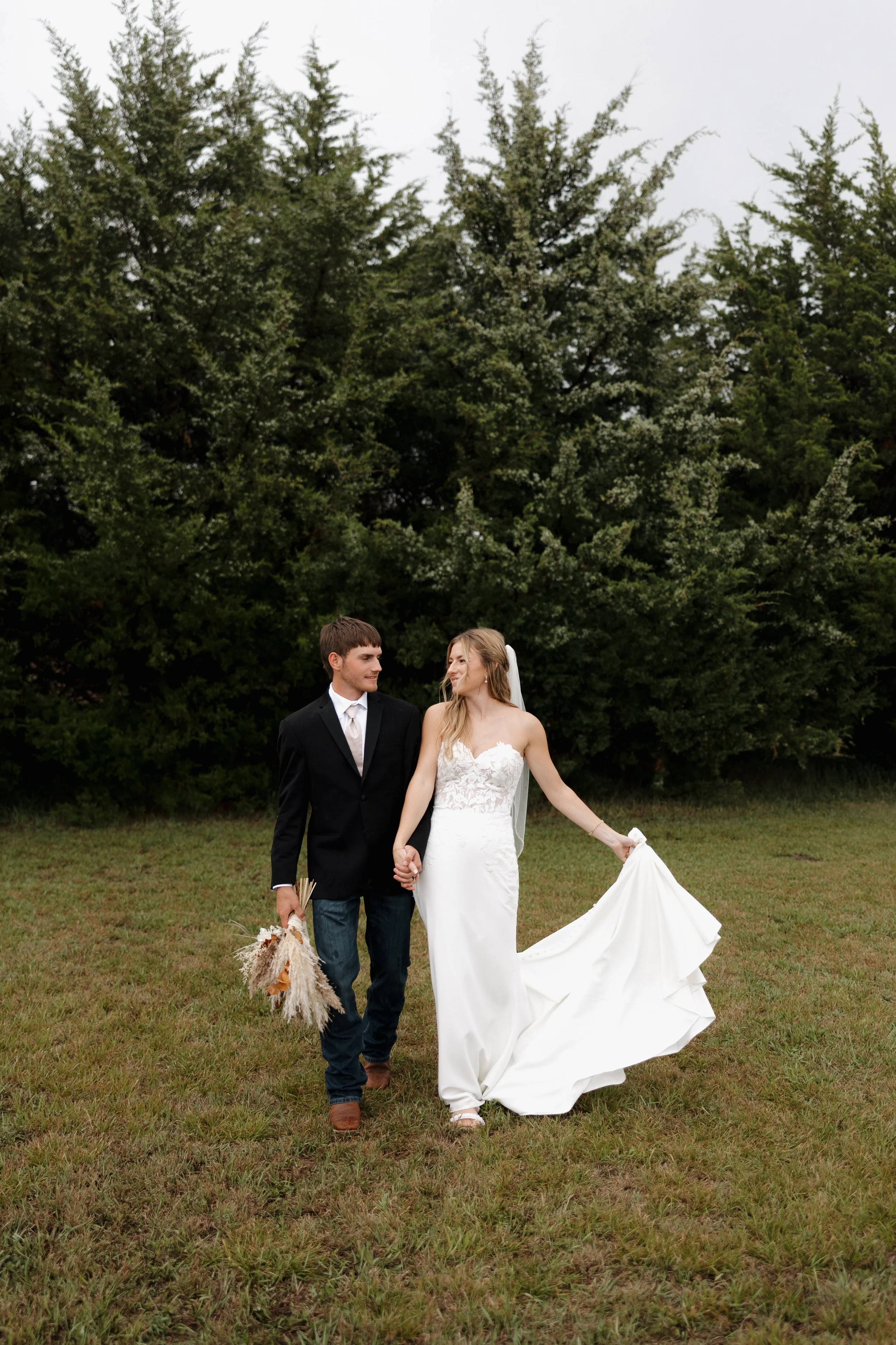 A bride and groom walking hand in hand outdoors on a grassy field with trees in the background, dressed in wedding attire; the bride in a white gown and the groom in a suit.