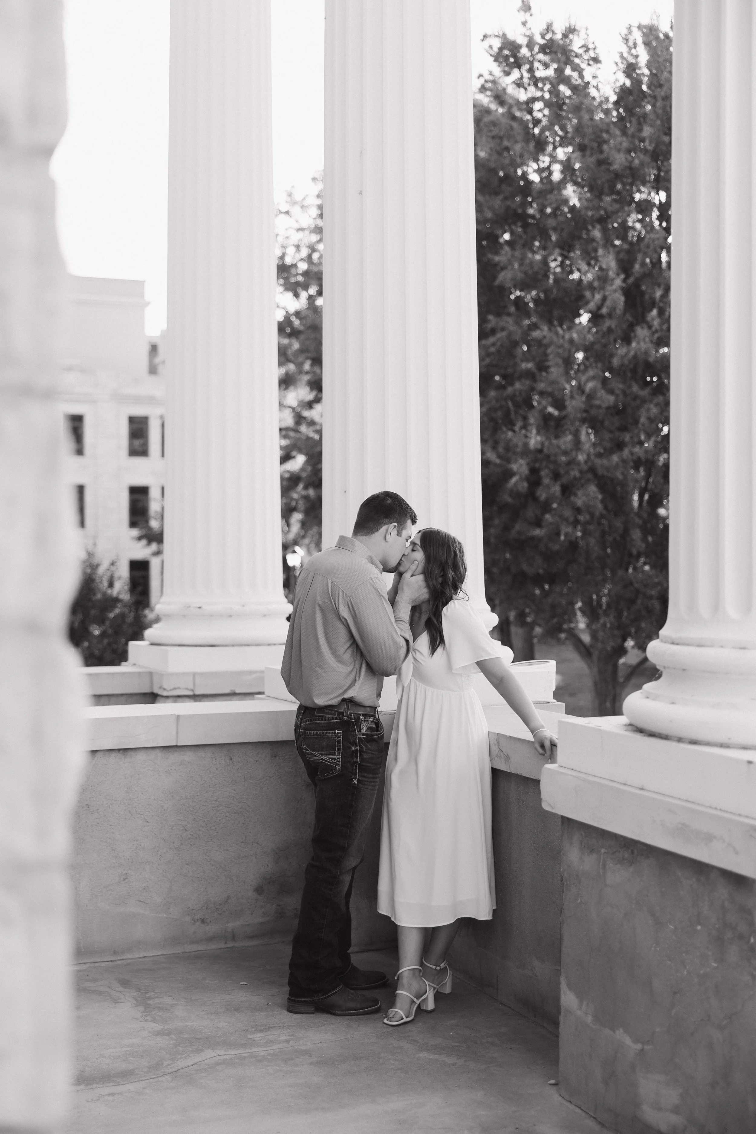 A couple is sharing an intimate moment on a porch with large white columns, touching foreheads. The man is wearing a button-up shirt and jeans, while the woman is dressed in a flowing white dress with high-heeled sandals. Trees and a building can be seen in the background.