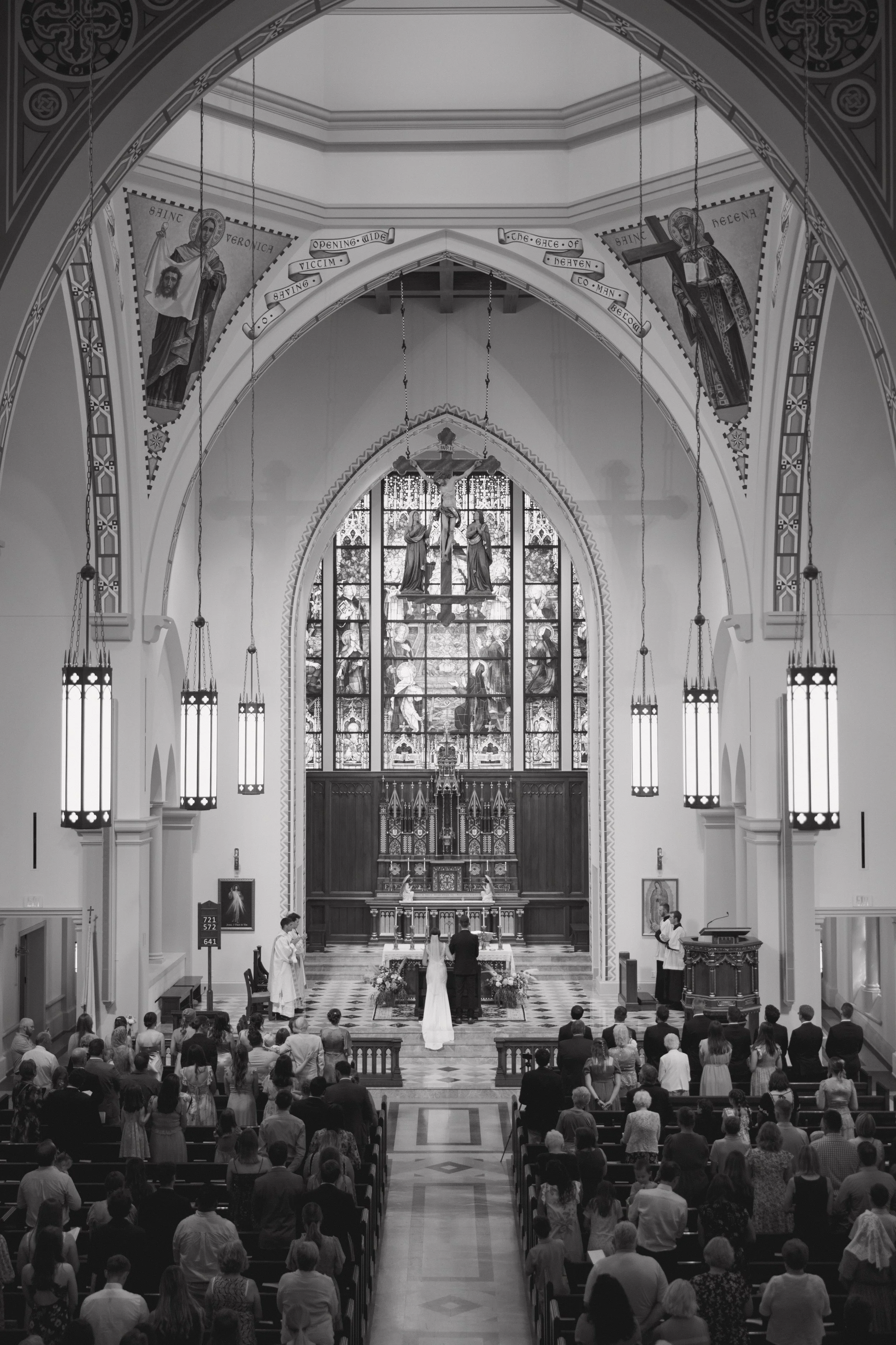 Interior of a church during a wedding ceremony with a congregation seated and standing, an altar at the front with a priest and a couple, stained glass windows behind the altar, and religious artwork and banners hanging from the ceiling.