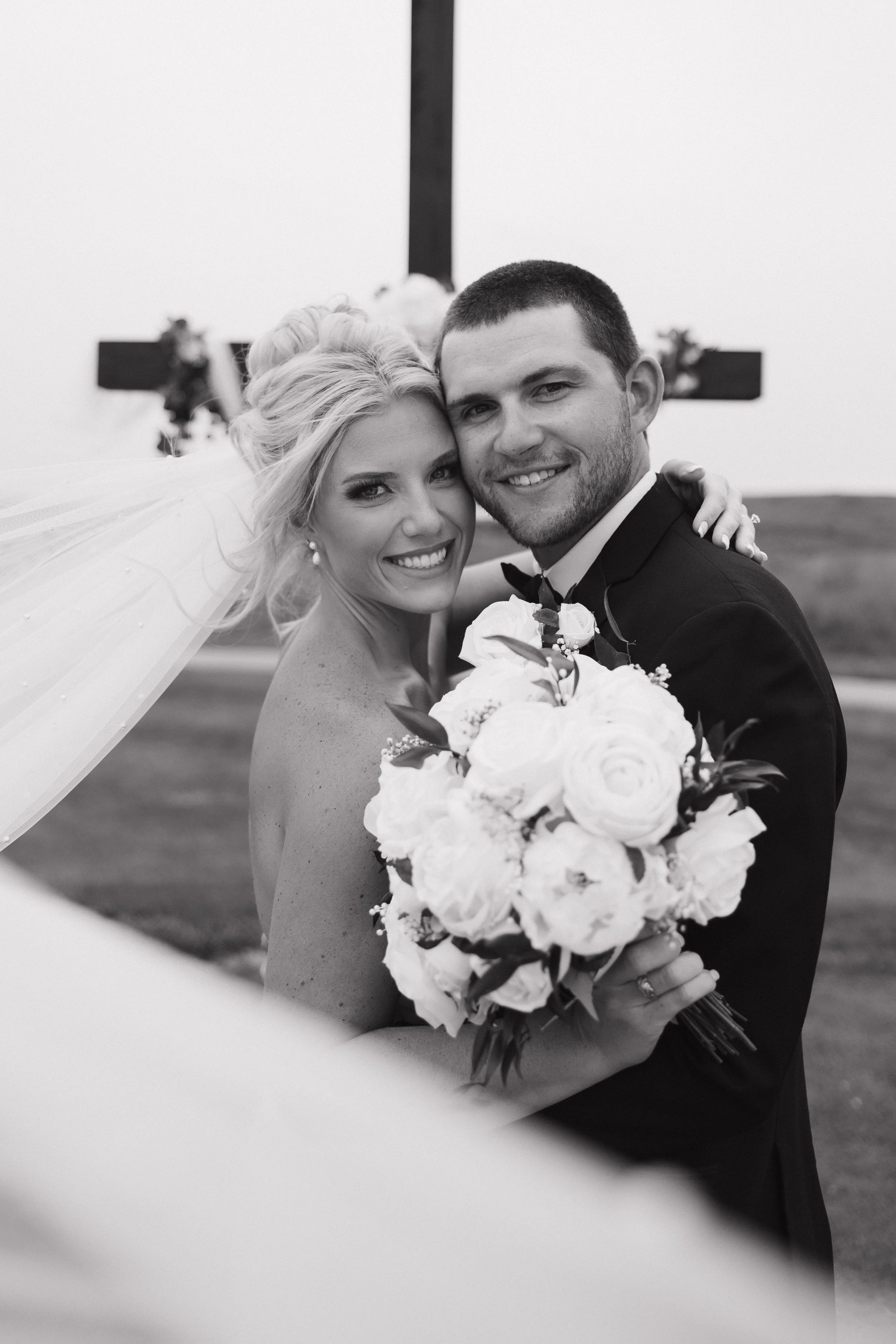 A black and white photo of a bride and groom smiling and embracing in front of a large cross, holding a bouquet of flowers, outdoors.