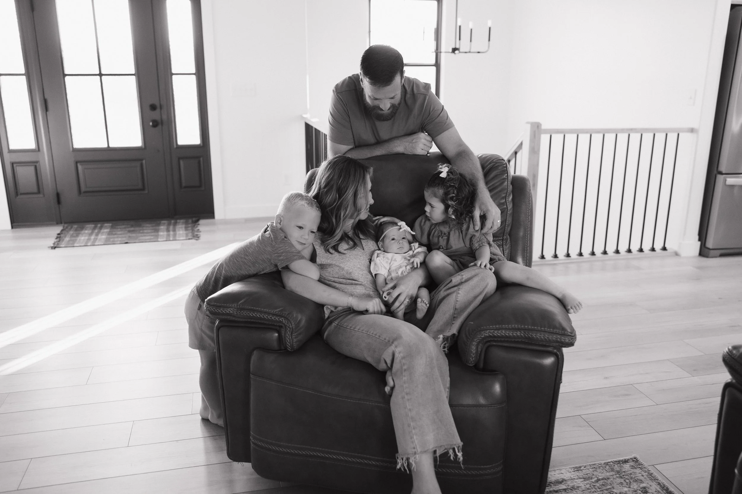A black and white photo of a family gathered on a sofa in a well-lit living room. The mother is sitting on the couch holding a baby, with an older child sitting on her lap. A man, likely the father, is leaning over from behind the sofa, interacting w