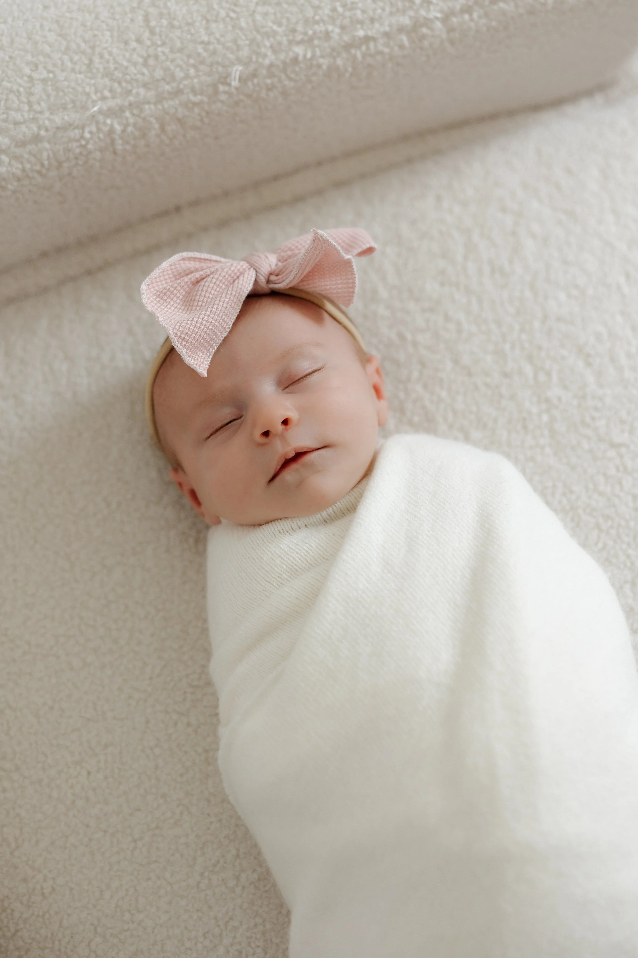 Close-up photo of a sleeping newborn baby wearing a pink bow headband and wrapped in a white blanket, lying on a beige textured surface.