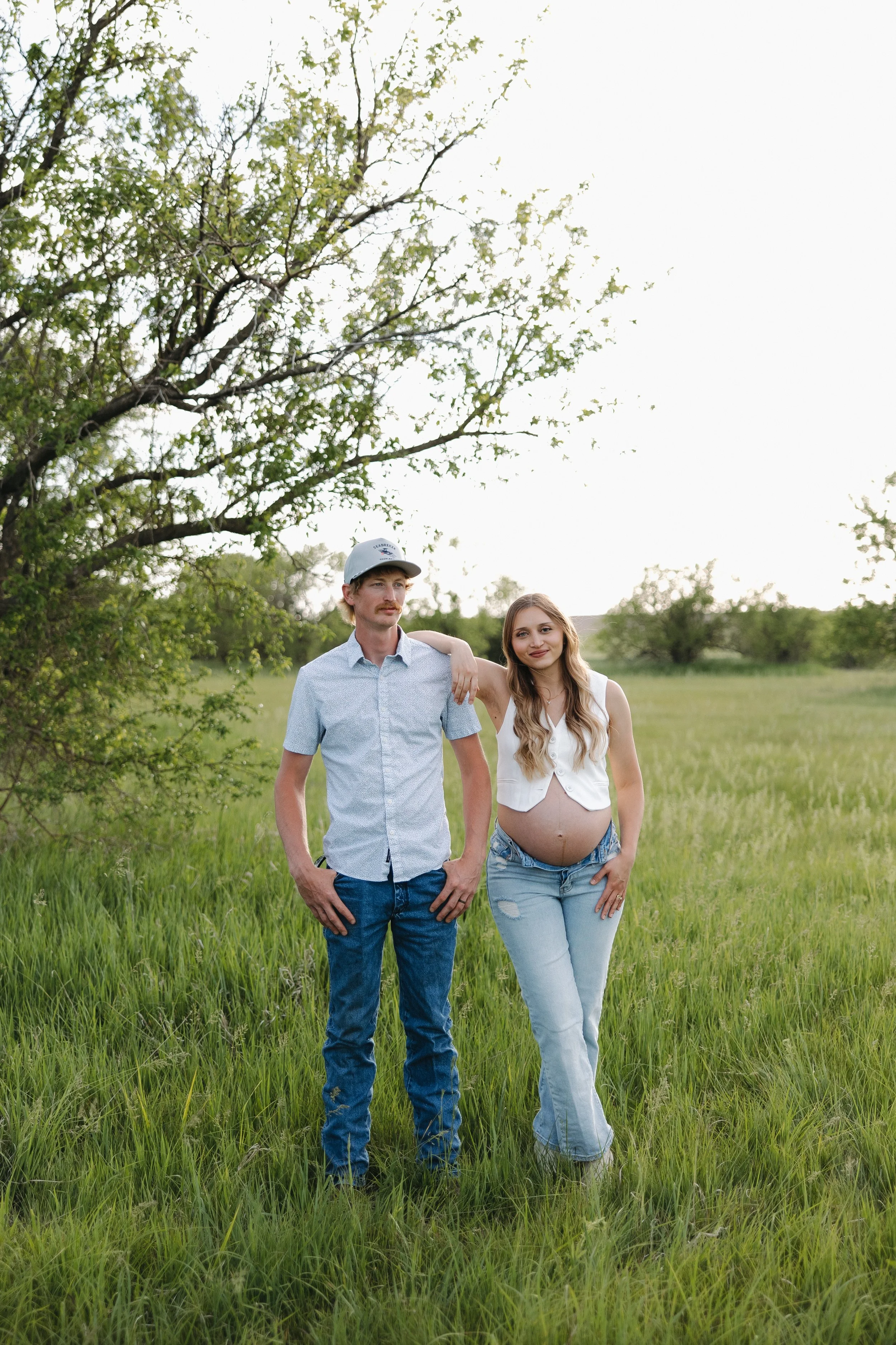A pregnant woman with long blonde hair wearing a white sleeveless top and ripped jeans, standing next to a man with a mustache wearing a light blue button-up shirt, a cap, and jeans, in a grassy field with trees in the background.