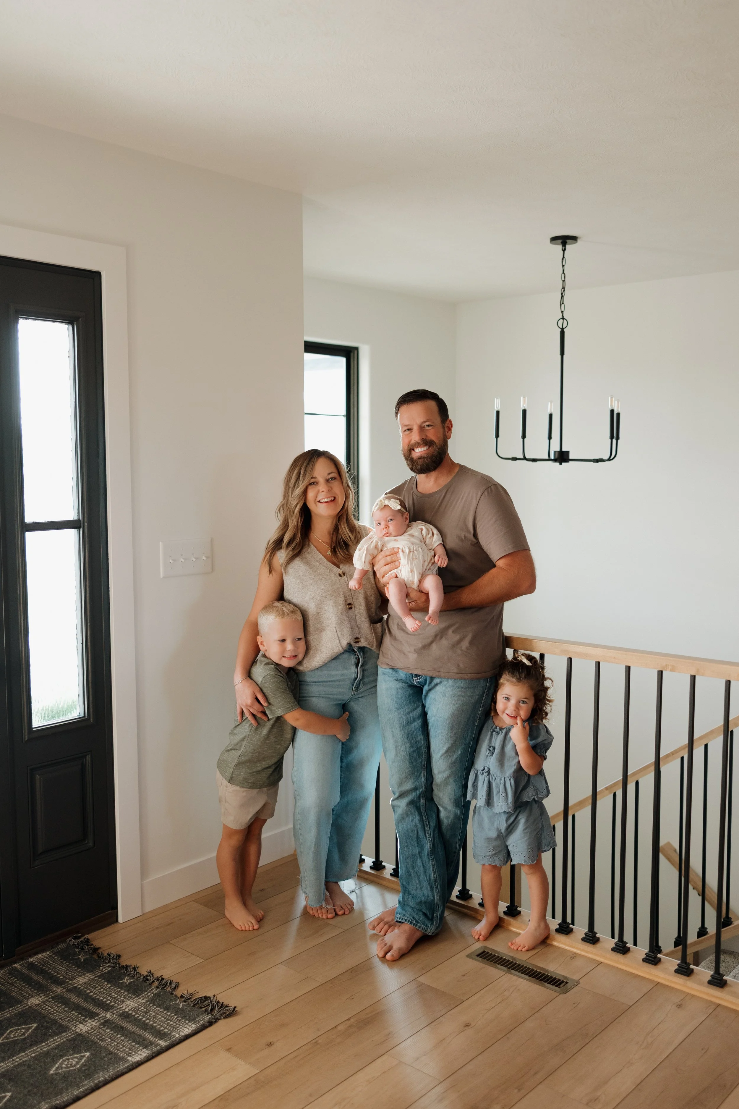 A family of six standing on a wooden floor in a home, smiling and posing for the photo. The father is holding a baby girl, and there are two young children standing beside and in front of them, with a woman and a man in casual clothing.