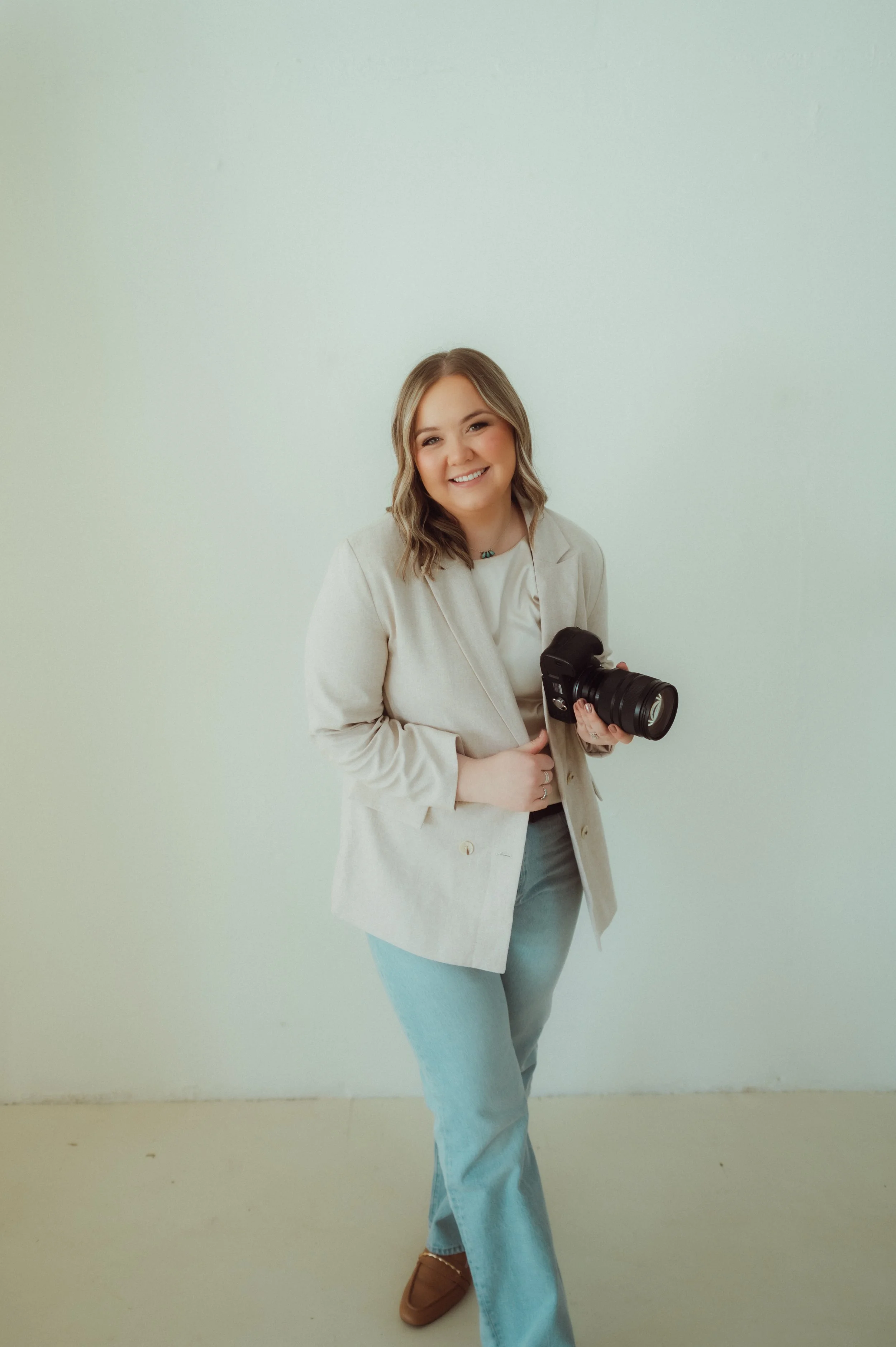 A smiling young woman with wavy light brown hair holding a camera, wearing a beige blazer, light-colored top, light blue jeans, and brown shoes, standing against a plain light green wall.