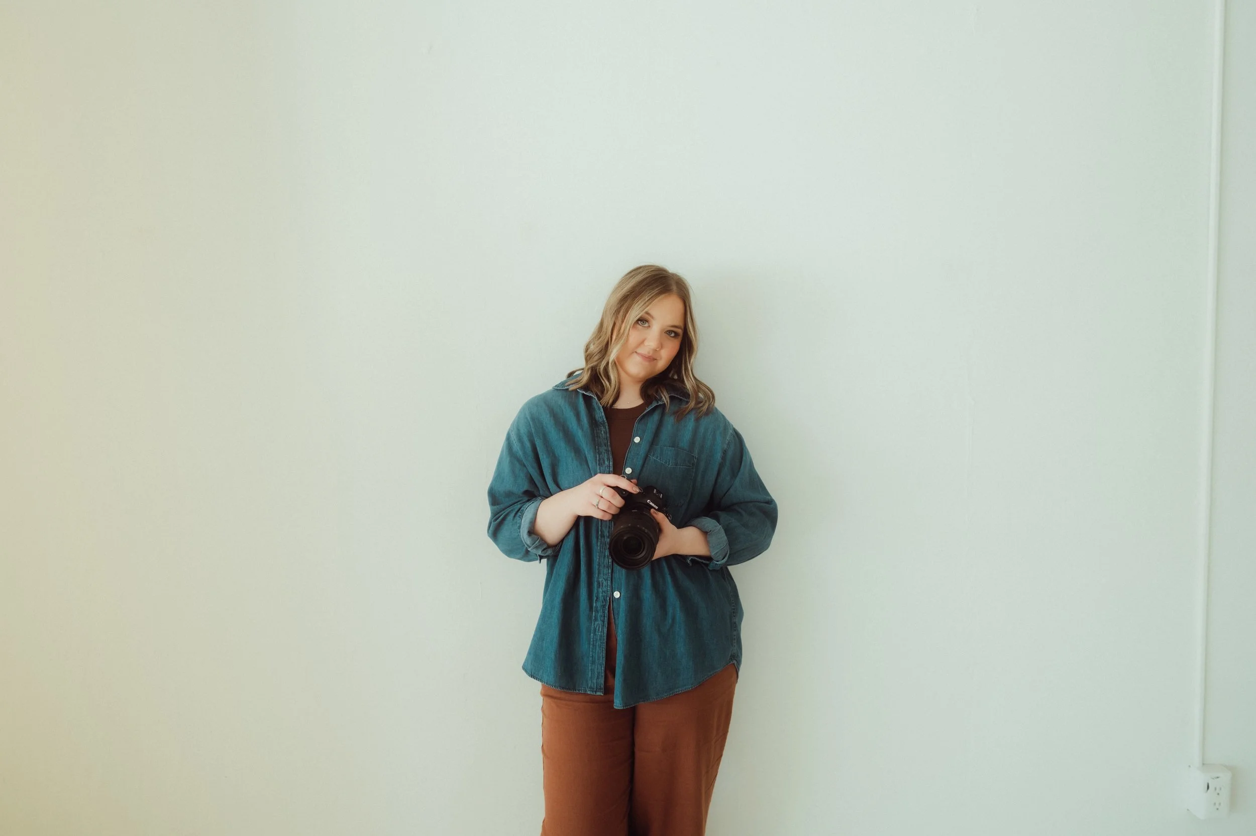 A woman with wavy blonde hair wearing a denim shirt holding a camera, standing against a plain white wall.
