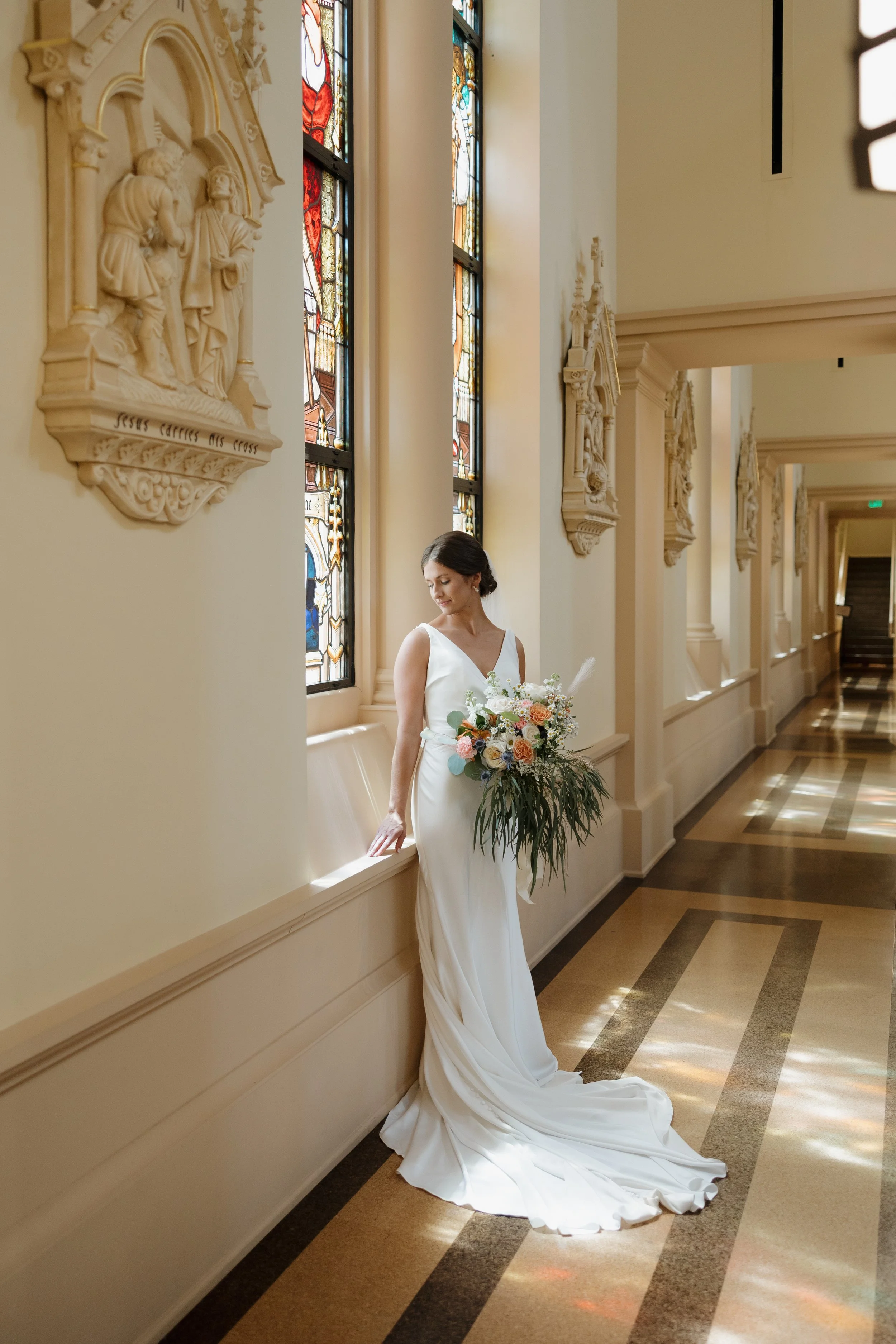 A bride in a white wedding dress holding a bouquet of flowers, standing in a corridor with decorative wall carvings and stained glass windows.