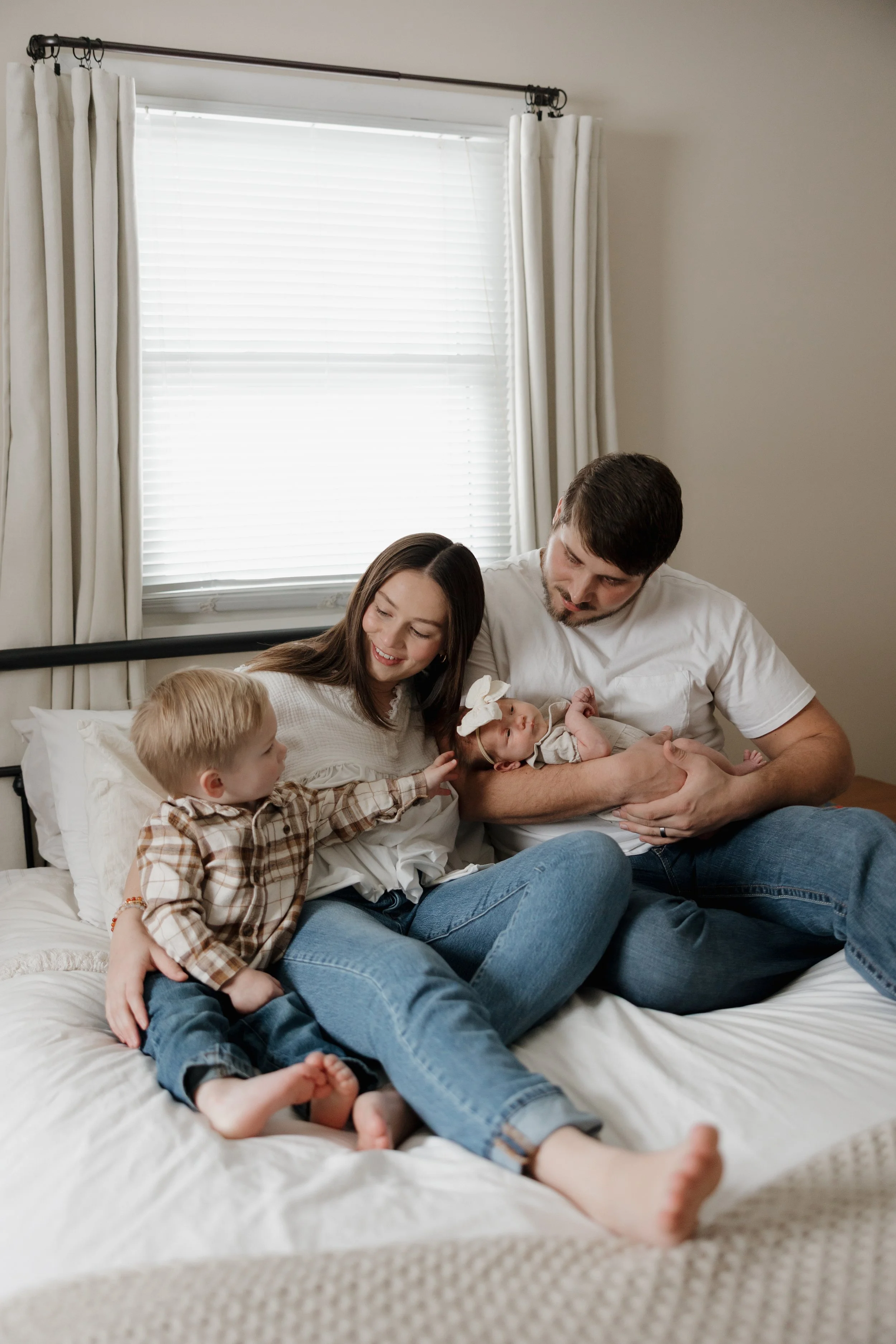 A family of five sitting on a bed in a bedroom, with a mother, father, two young children, and a newborn baby. The parents are holding the baby, and the children are interacting with the baby and each other. The room has a large window with white bli