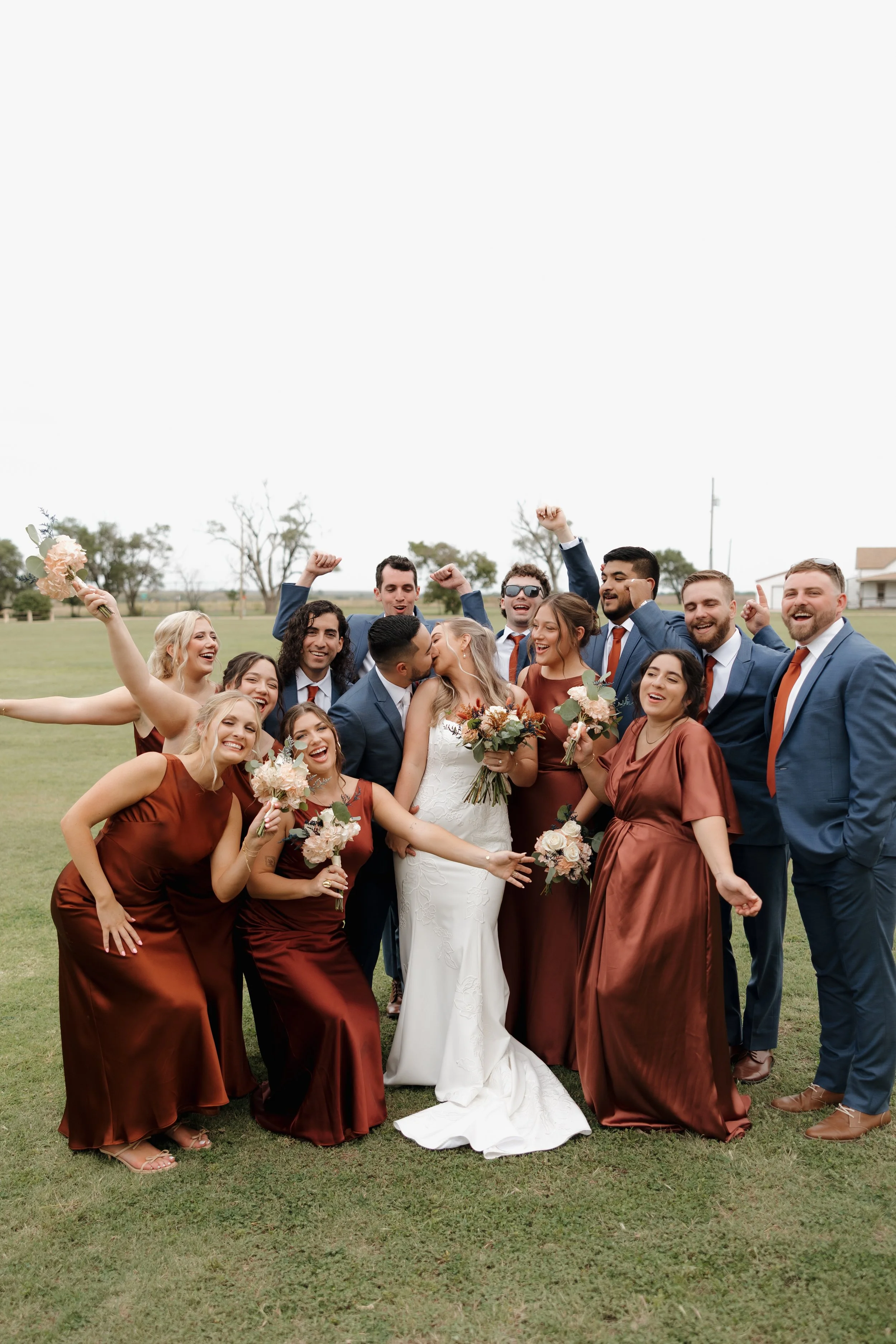 A joyful wedding group photo outdoors with the bride and groom in the center, surrounded by bridesmaids and groomsmen celebrating and smiling, with some raising their fists and holding bouquets.