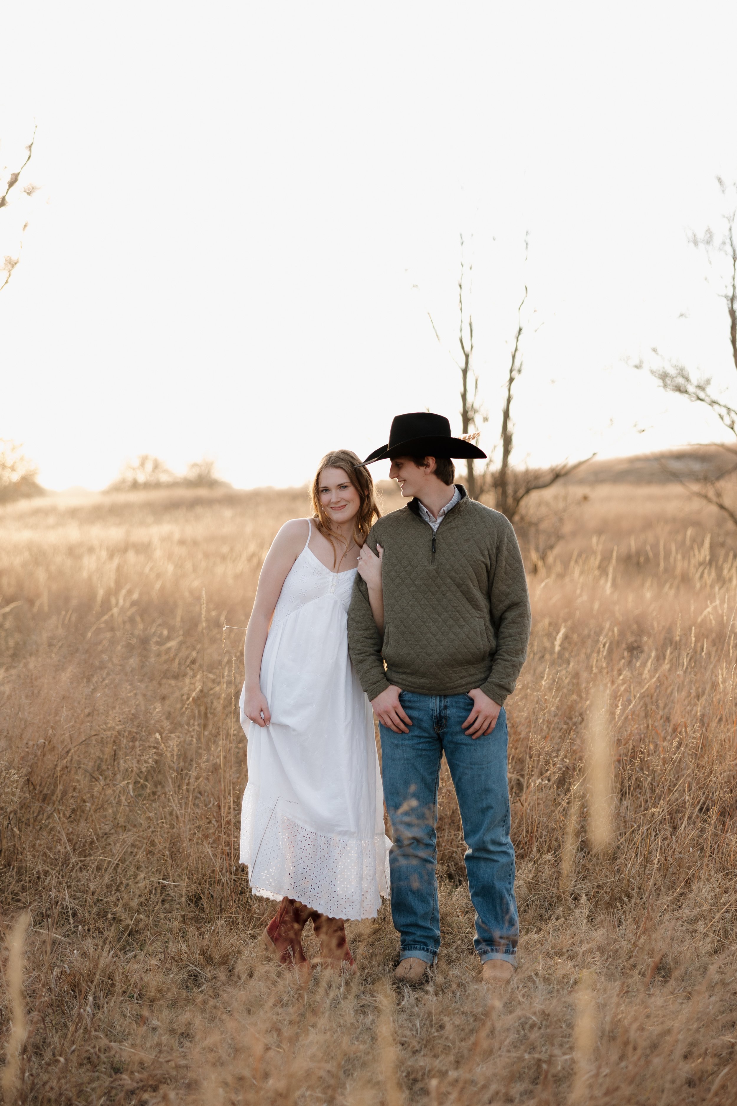 A man and woman standing together in a field during sunset, with the woman dressed in a white dress and the man wearing a cowboy hat and green sweater.