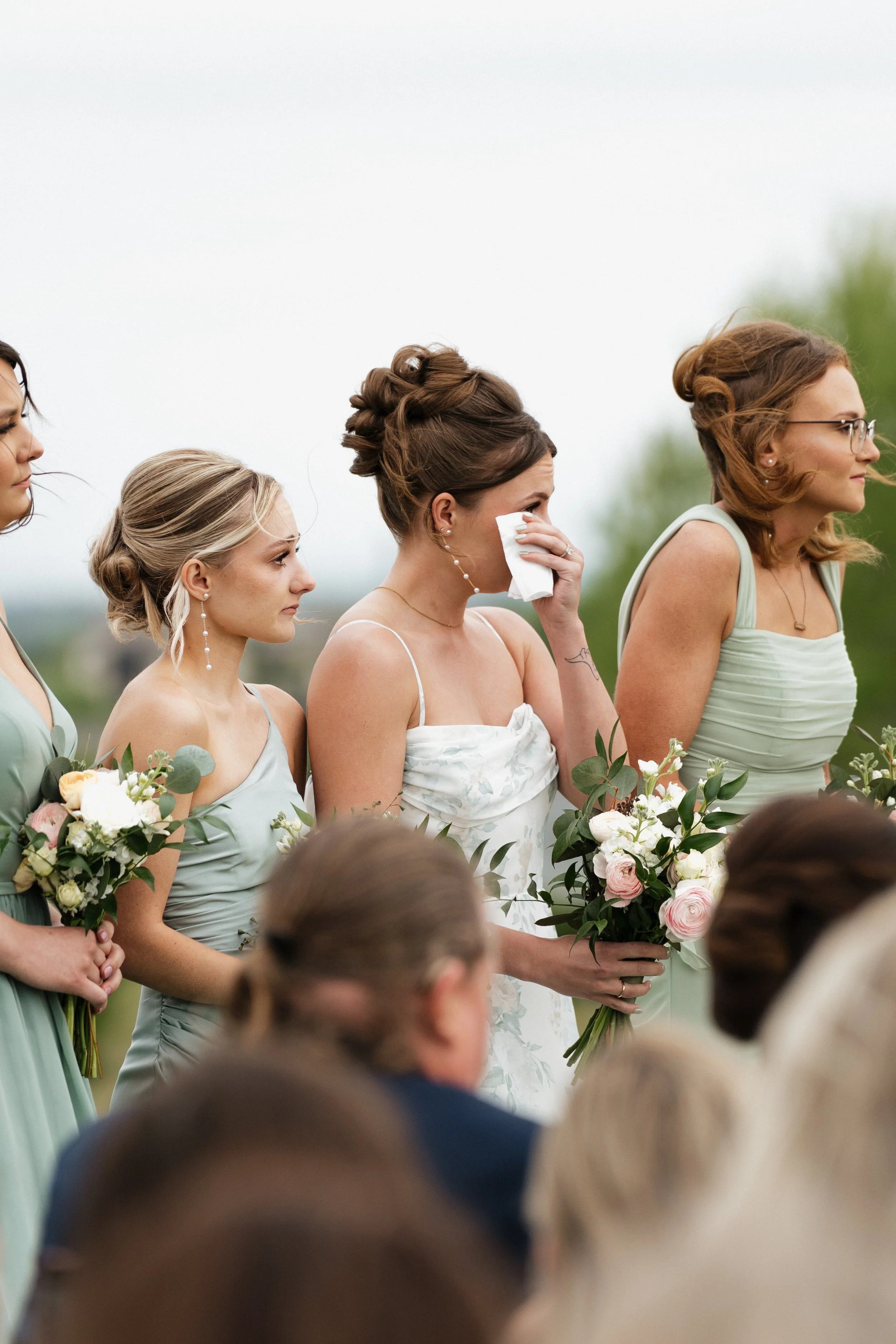 A group of women dressed in pastel-colored dresses, holding bouquets of flowers, standing outdoors on a cloudy day during a wedding ceremony.