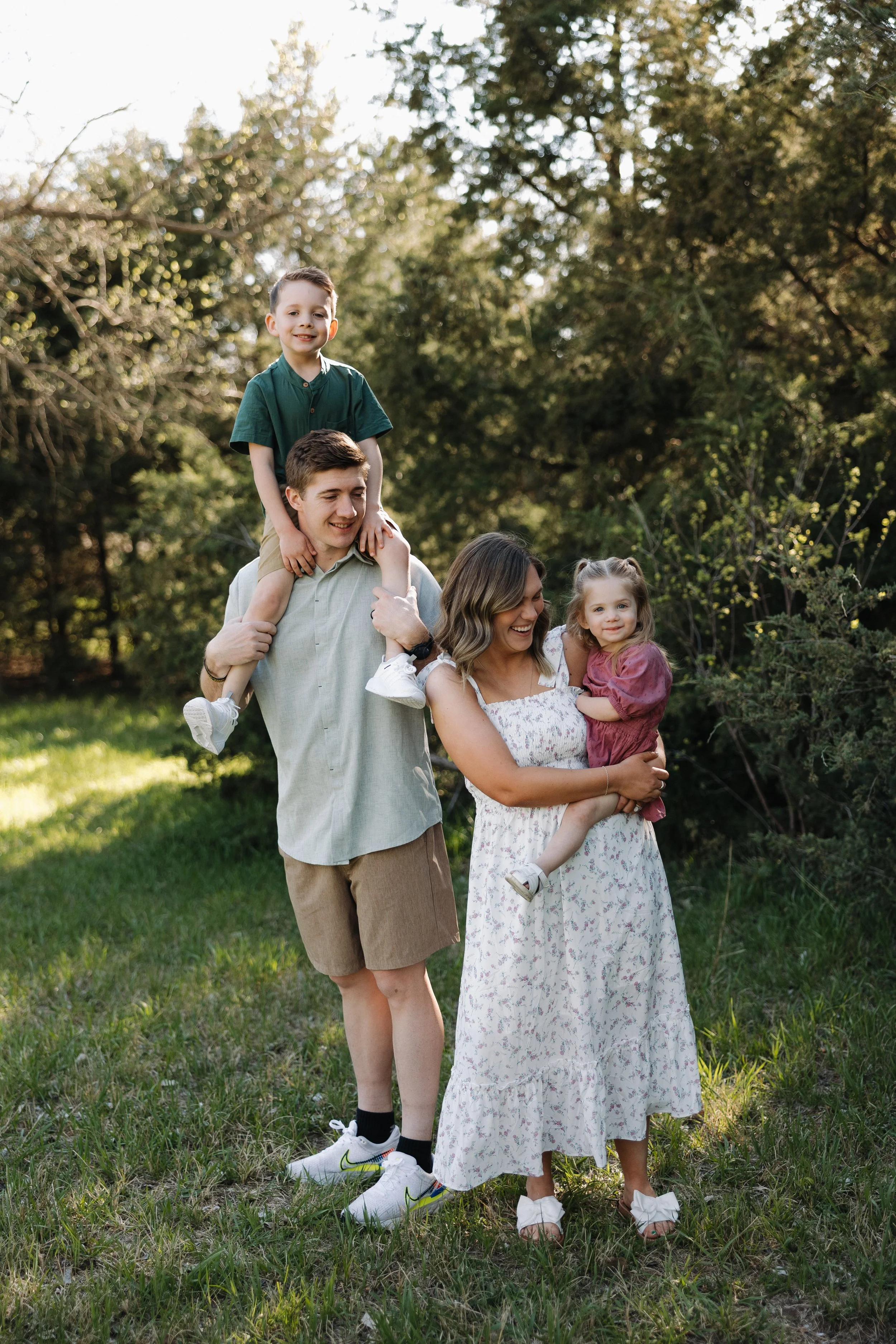 Family outdoors in a park with trees, a man carrying a boy on his shoulders, and a woman holding a girl in her arms, smiling and enjoying the sunshine.
