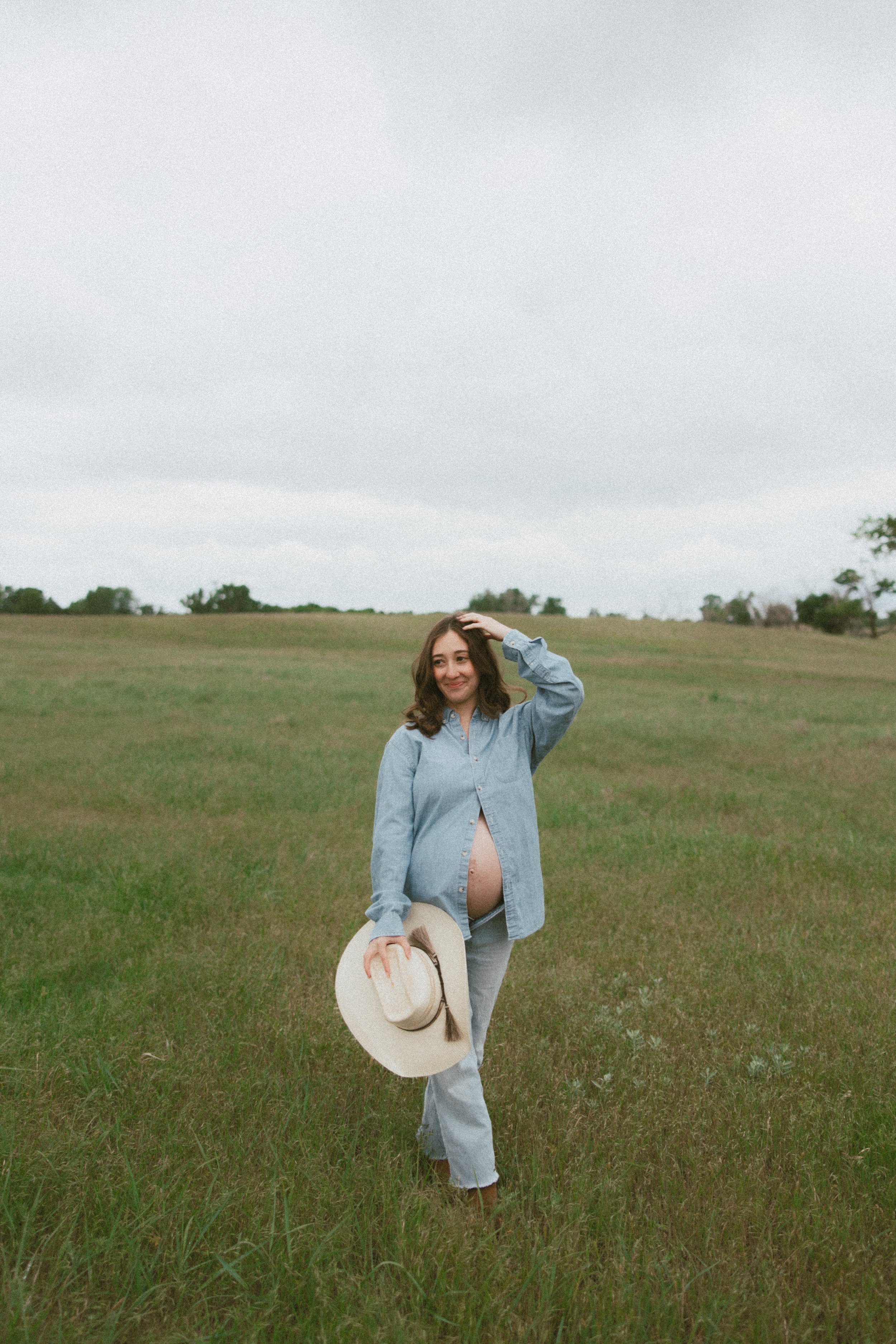 A pregnant woman standing in a grassy field holding a hat and wearing an unbuttoned denim shirt, smiling and has her hand on her head as she looks to the side.