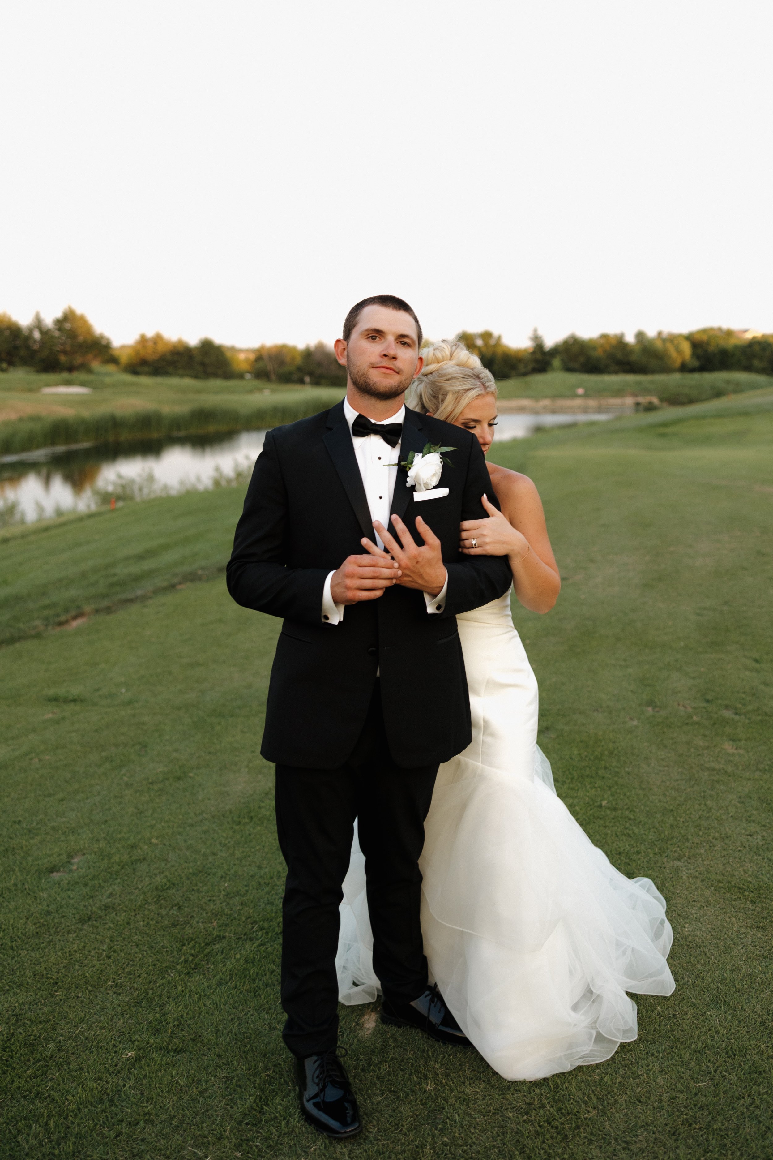 A man in a black tuxedo with a bow tie stands next to a woman in a white wedding gown with a full skirt. They are on a grassy area near a body of water with trees in the background. The man has his hands clasped in front, and the woman is slightly behind him, with her arm around his waist and her hand on his chest.