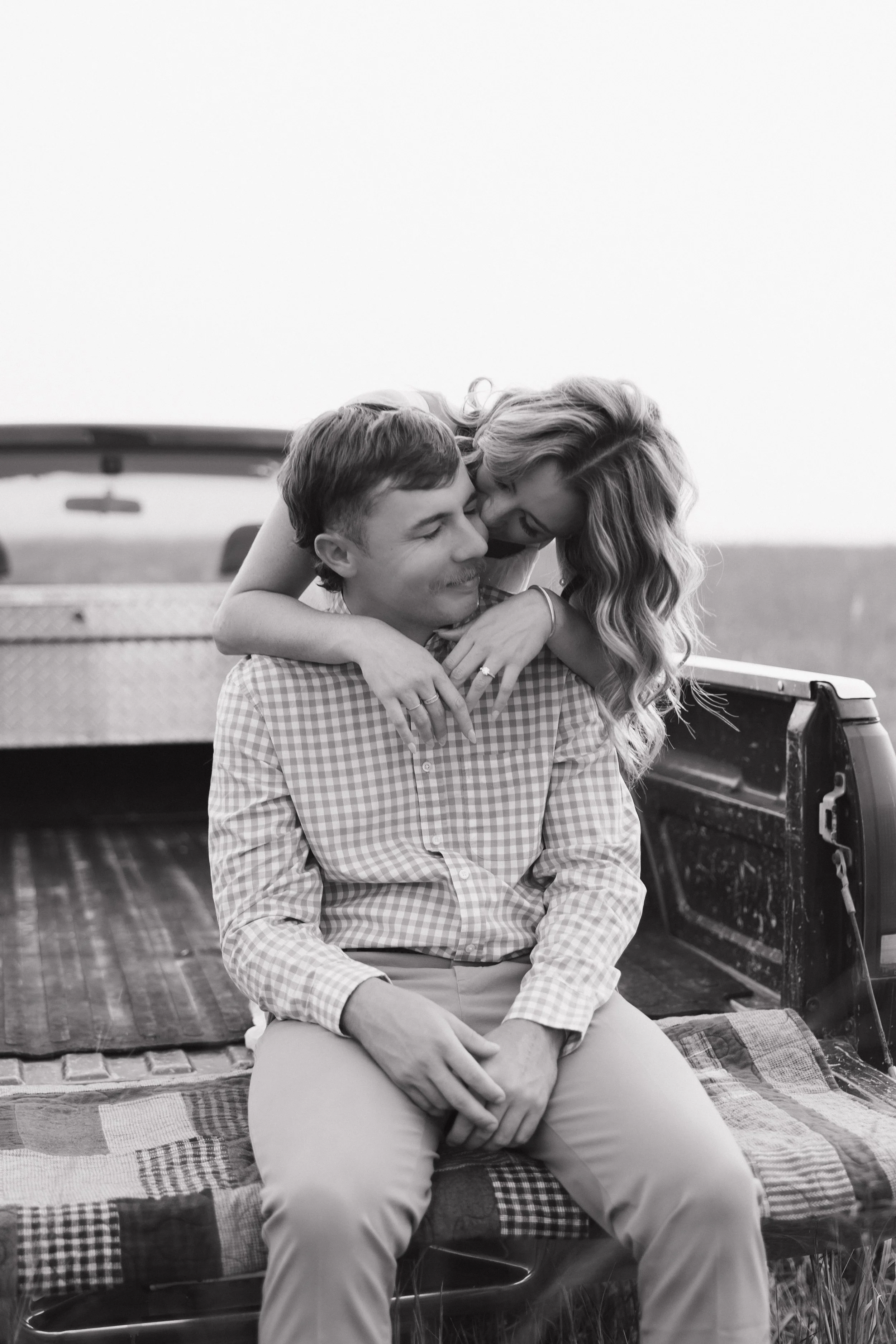 A man and woman sharing a hug in the back of a pickup truck, with a quilt on the truck bed, outdoors in a grassy field.