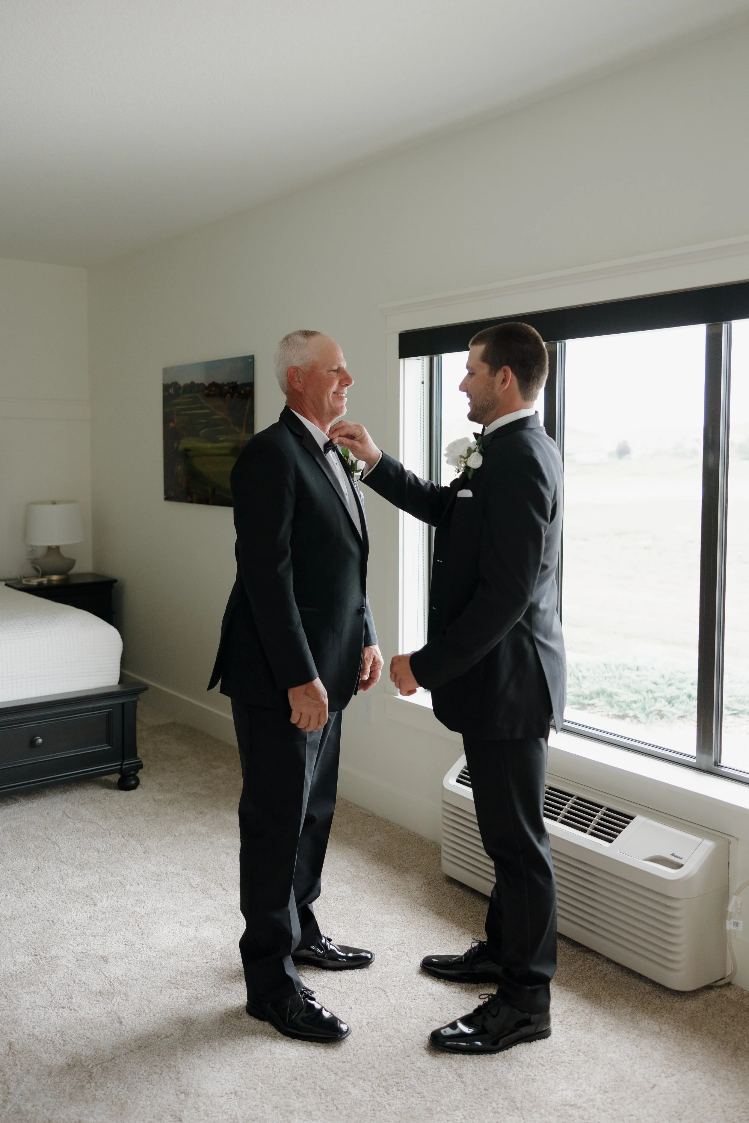 A father and son, both dressed in black tuxedos with white shirts and black bow ties, share a joyful moment in a bedroom as the father adjusts the son's boutonniere. They stand by a large window with a view of the outdoors, near an air conditioning u