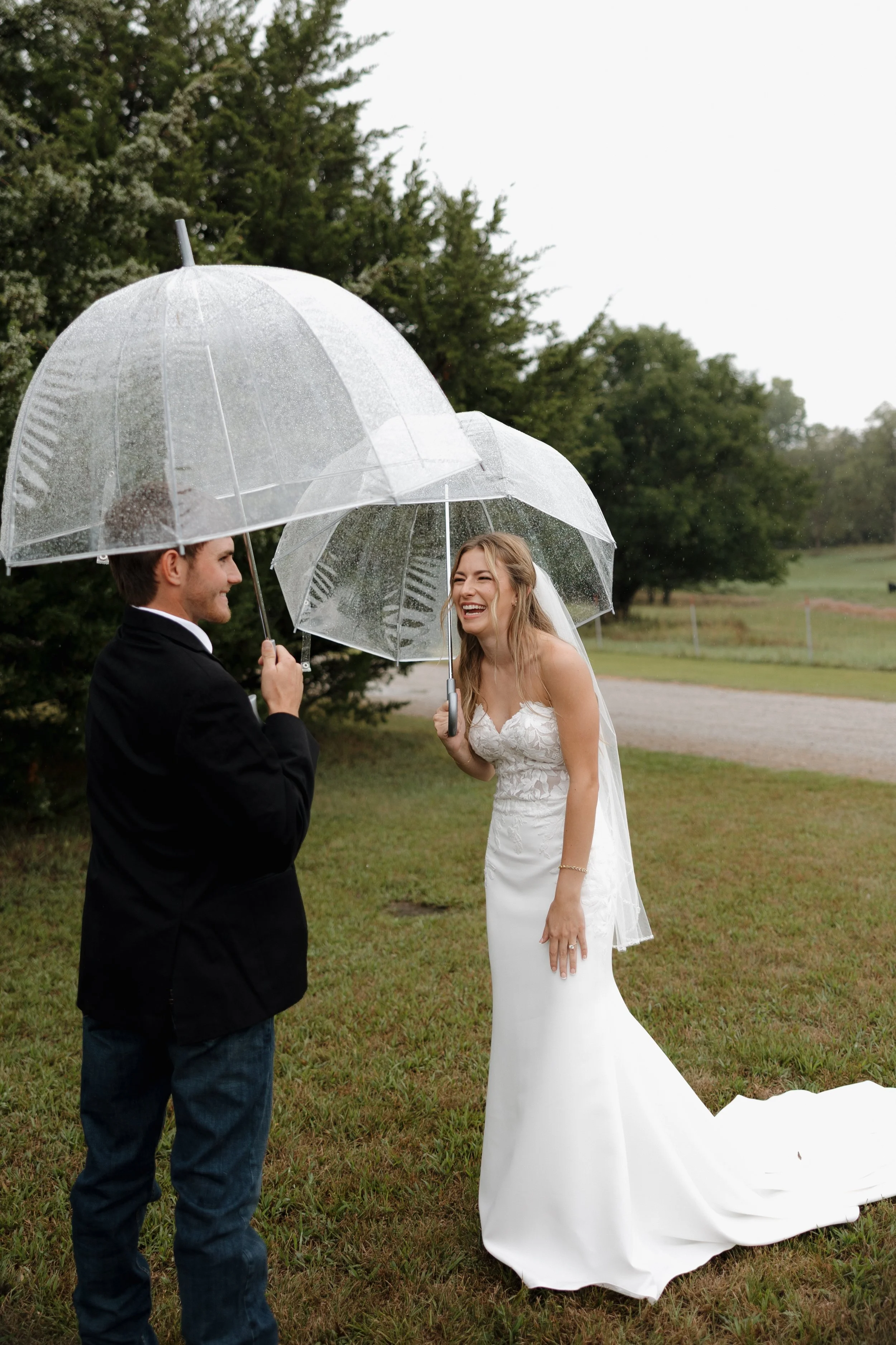 A bride and groom smiling and laughing under transparent umbrellas in the rain on their wedding day outside, with trees and grass around.