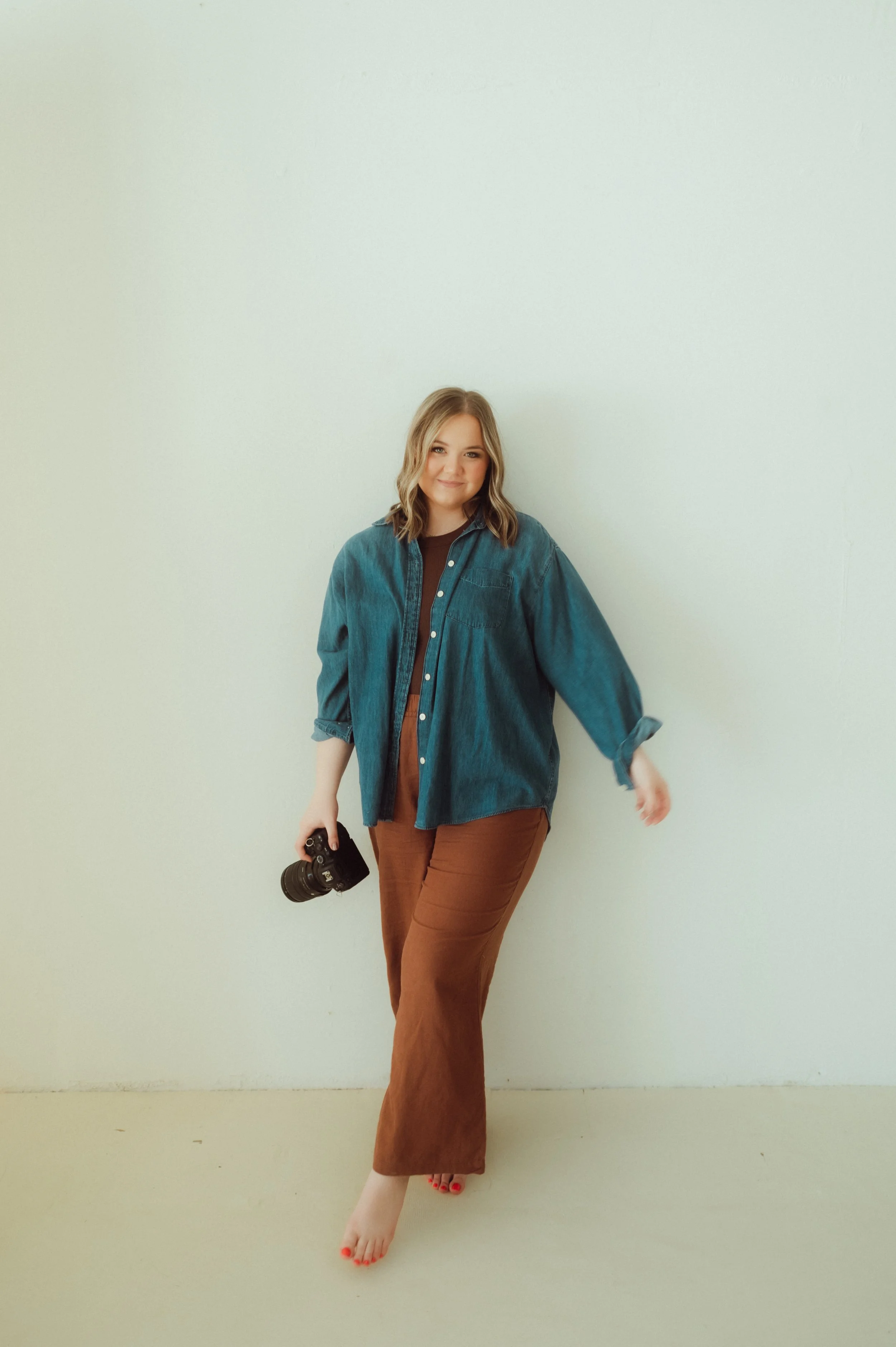 Young woman in a denim jacket and brown pants holding a camera stands barefoot against a plain white wall.