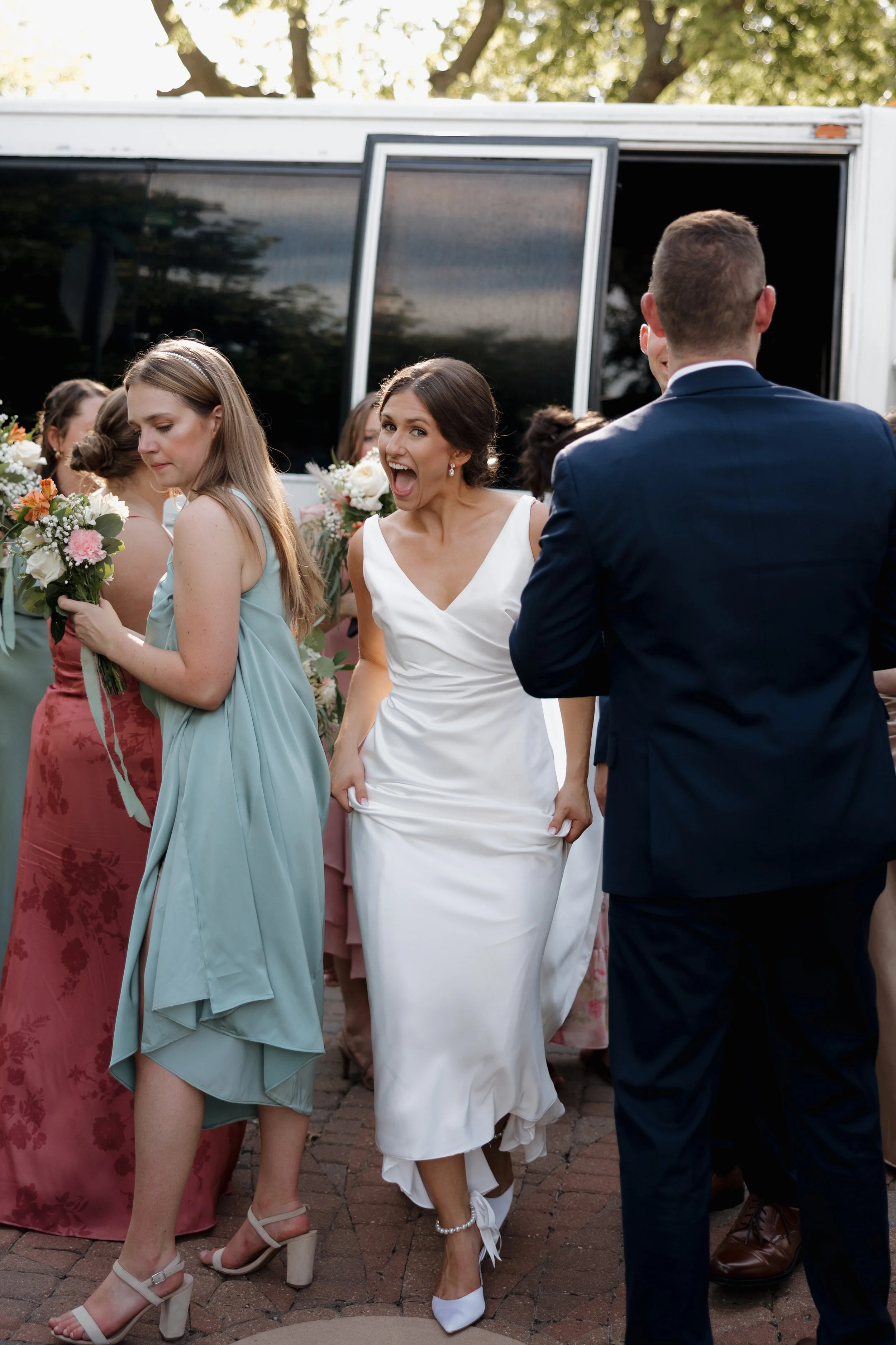 A group of people gathered outdoors, some dressed in formal attire. One woman in a white dress is smiling and appears excited, while others hold bouquets and wear pastel-colored dresses. The setting is sunny with trees in the background.