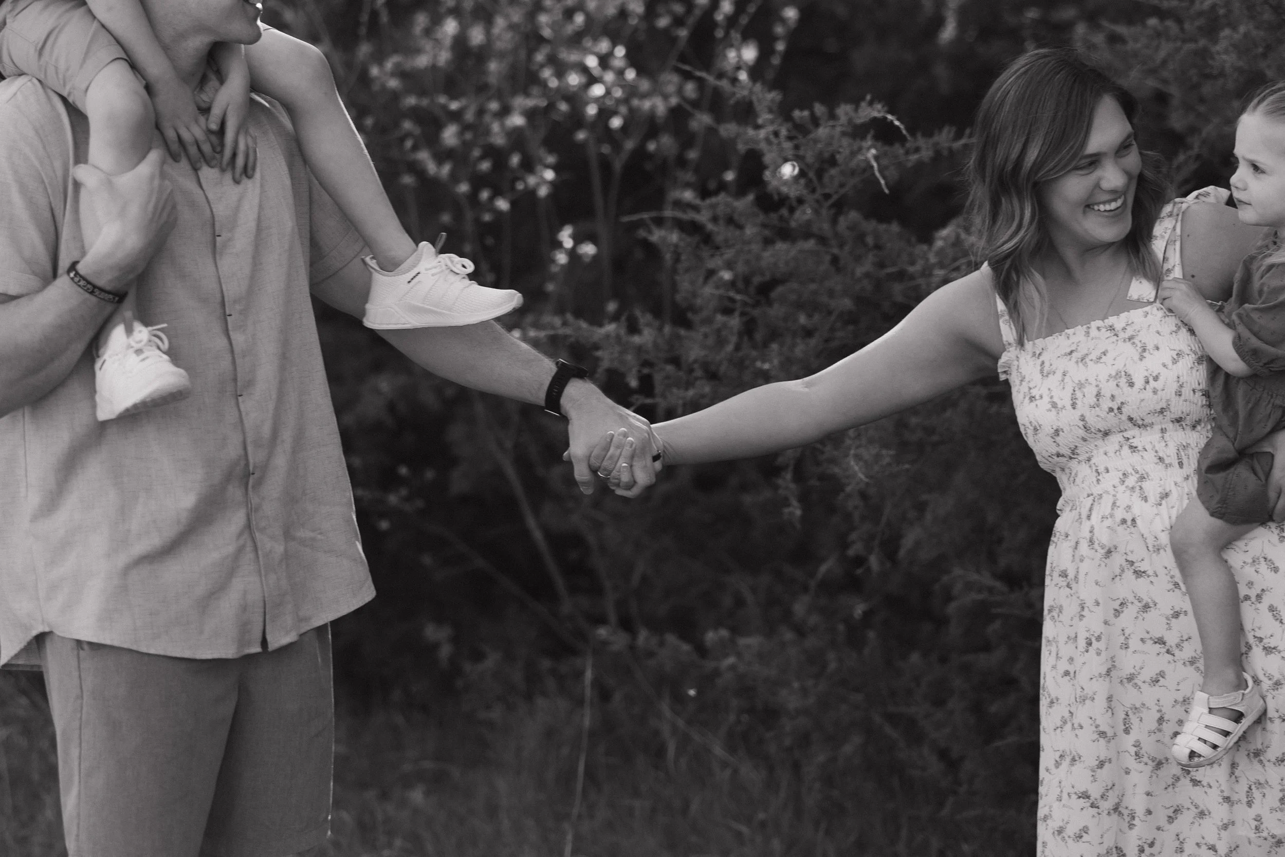 A woman holds hands with a child, while a man carries another child on his shoulders during an outdoor gathering in a park or garden. The scene is in black and white.