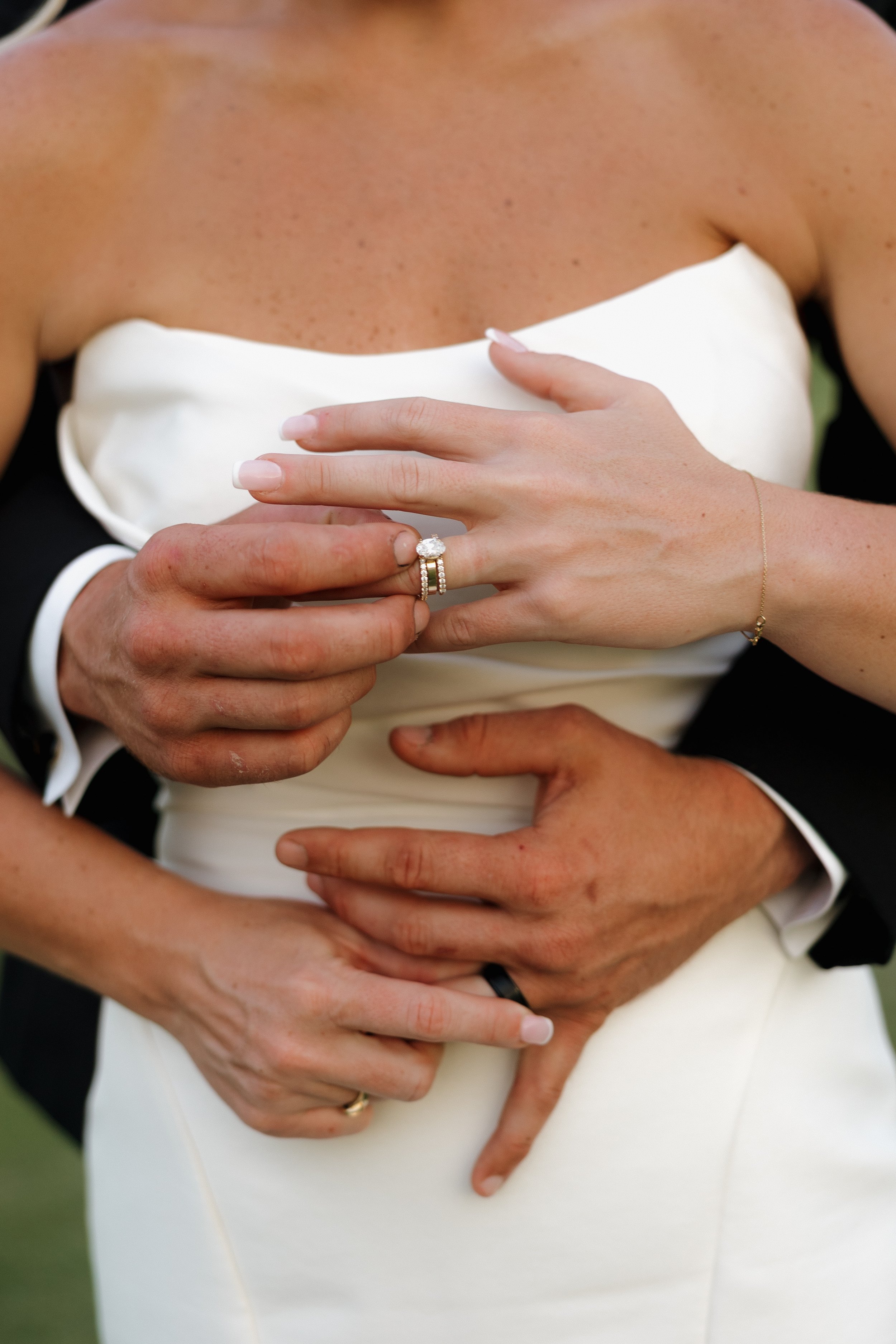 Close-up of a bride and groom embracing, with the bride wearing a white strapless dress and the groom in a black suit. The groom's hand is on the bride's waist, and the bride's hand is visible with a diamond engagement ring and wedding band. The brid