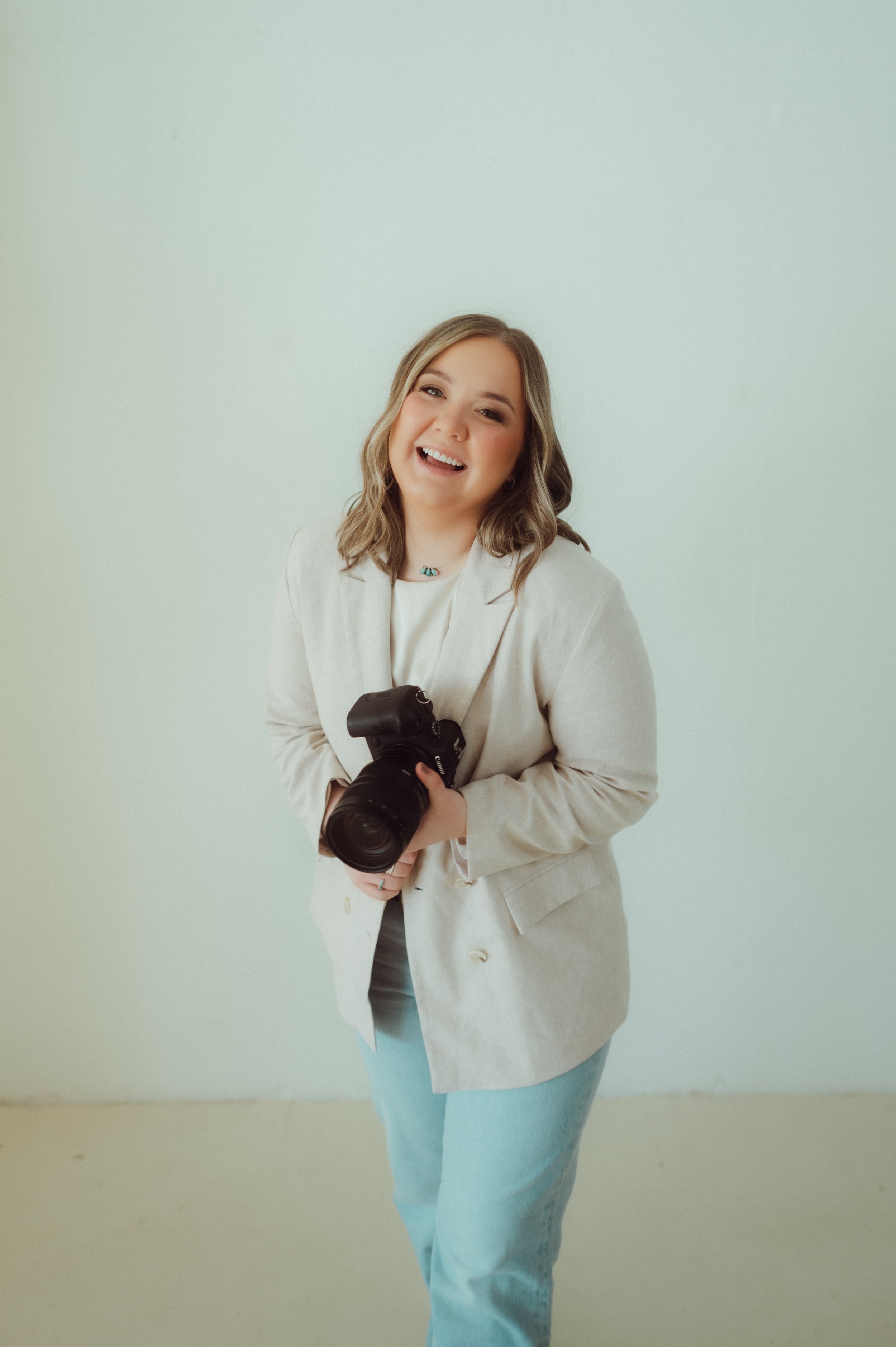 A woman smiling and holding a professional camera against a plain wall, wearing a beige blazer, light-colored top, and light blue jeans.
