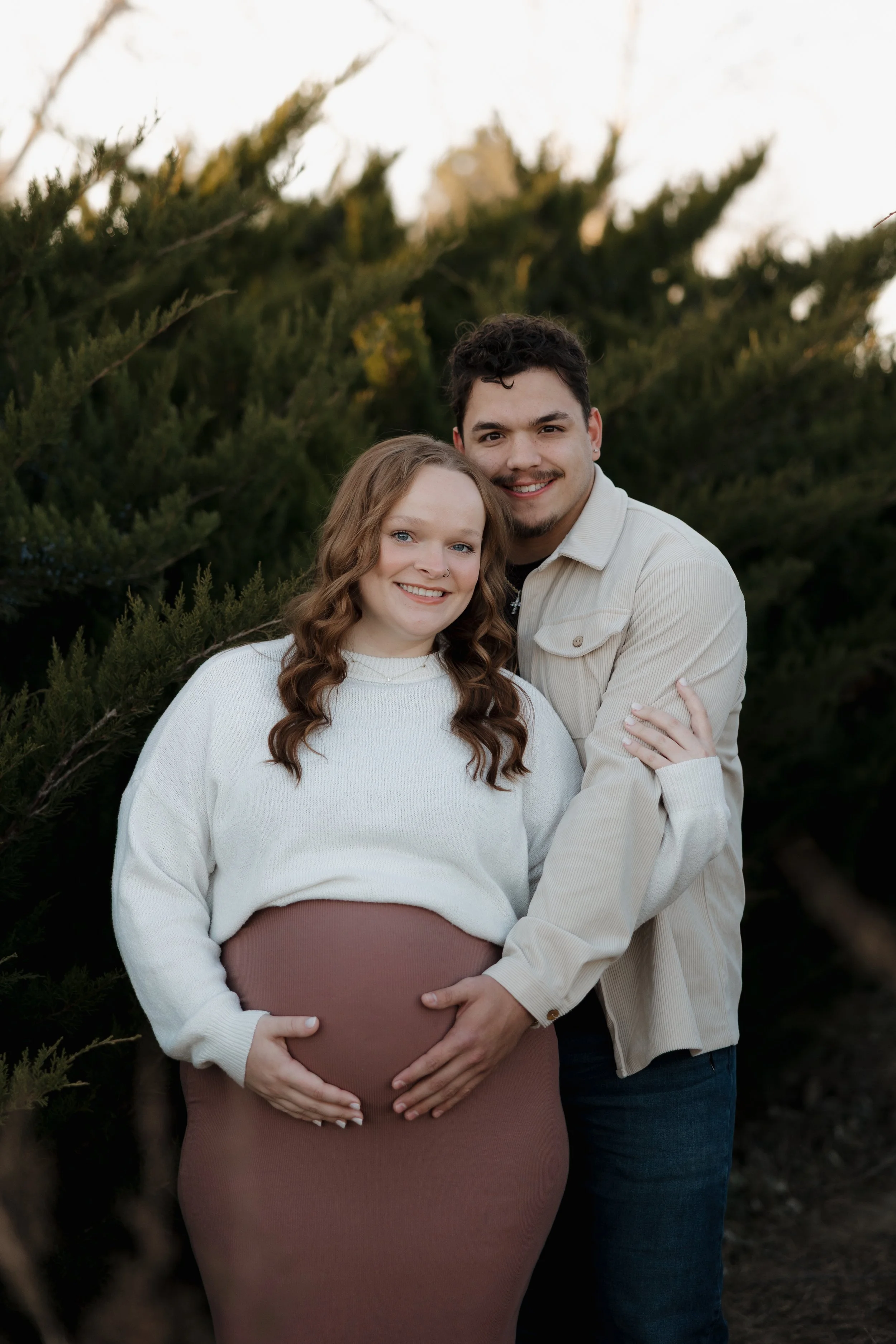 A pregnant woman with curly red hair wearing a white sweater and brown skirt standing outdoors surrounded by green bushes, with her partner, a man with dark hair and a mustache, wearing a light-colored jacket over a black shirt, standing behind her with his arms around her shoulders, both smiling at the camera.