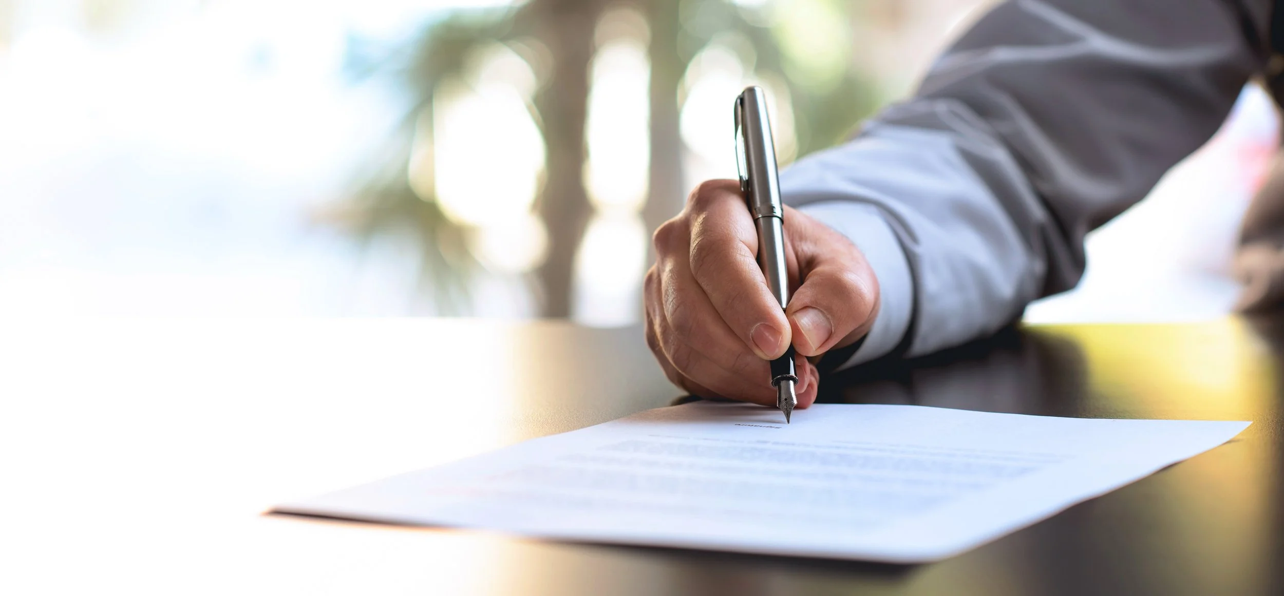 A person in a dress shirt writes with a pen on a sheet of paper as sunlight filters through trees in the background.