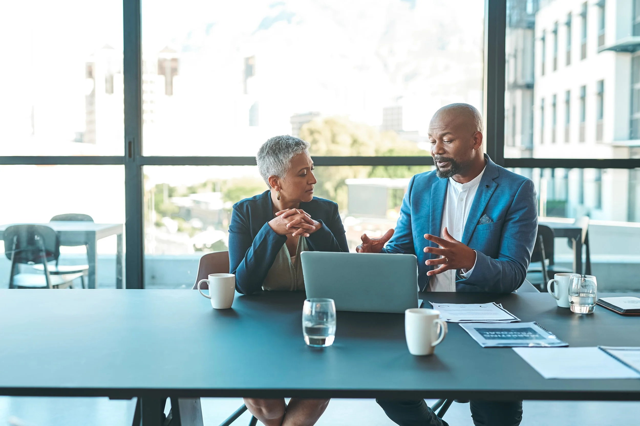 Two professionals engaged in a discussion during a meeting in a bright office with floor-to-ceiling windows. The woman, with short gray hair, is listening attentively, while the man, bald with a beard, is speaking and gesturing with his hands. On the table, there are a laptop, papers, a pen, and three white coffee mugs.