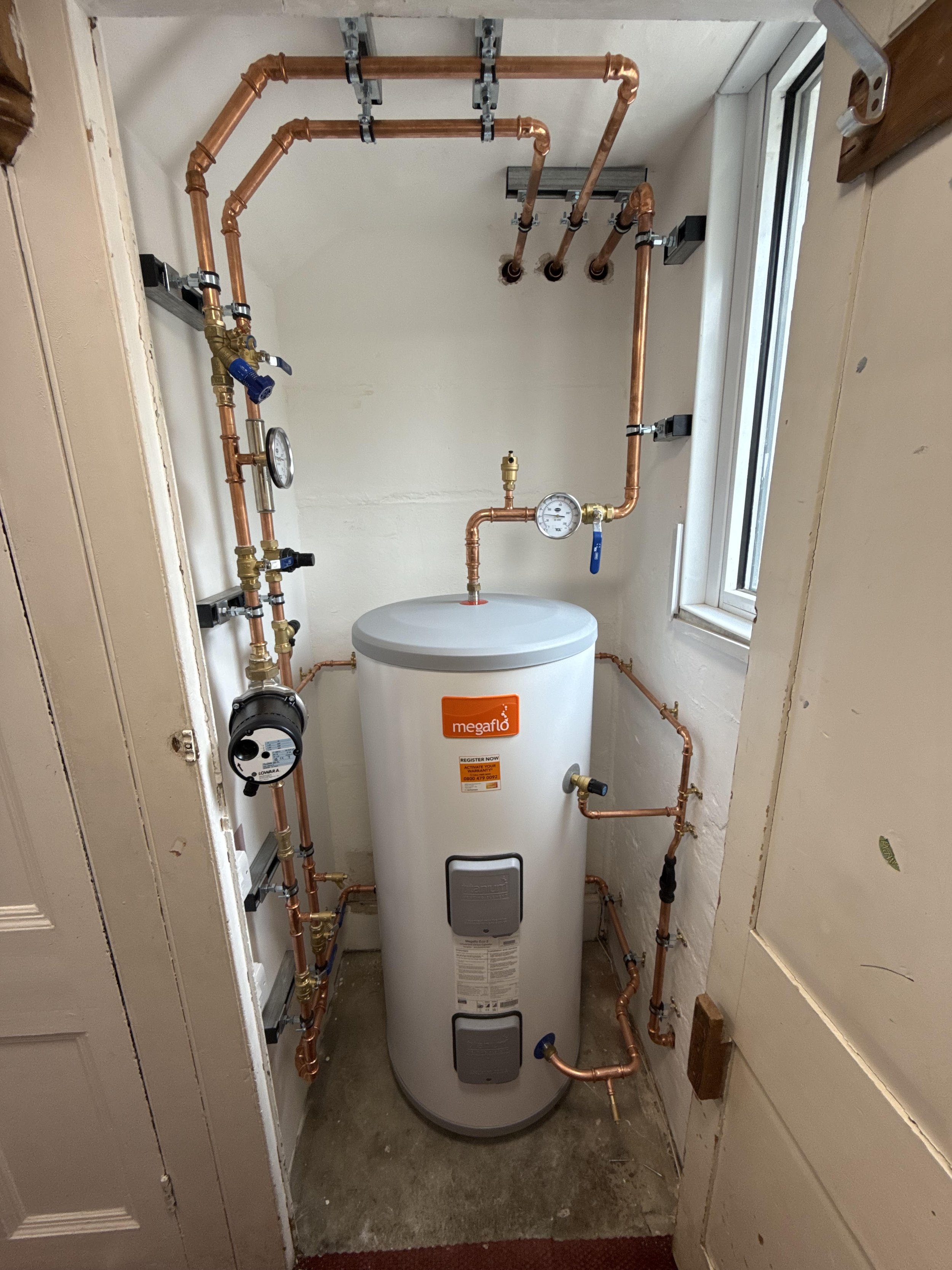 A water heater with copper plumbing pipes connected, installed in a small utility closet with a window.