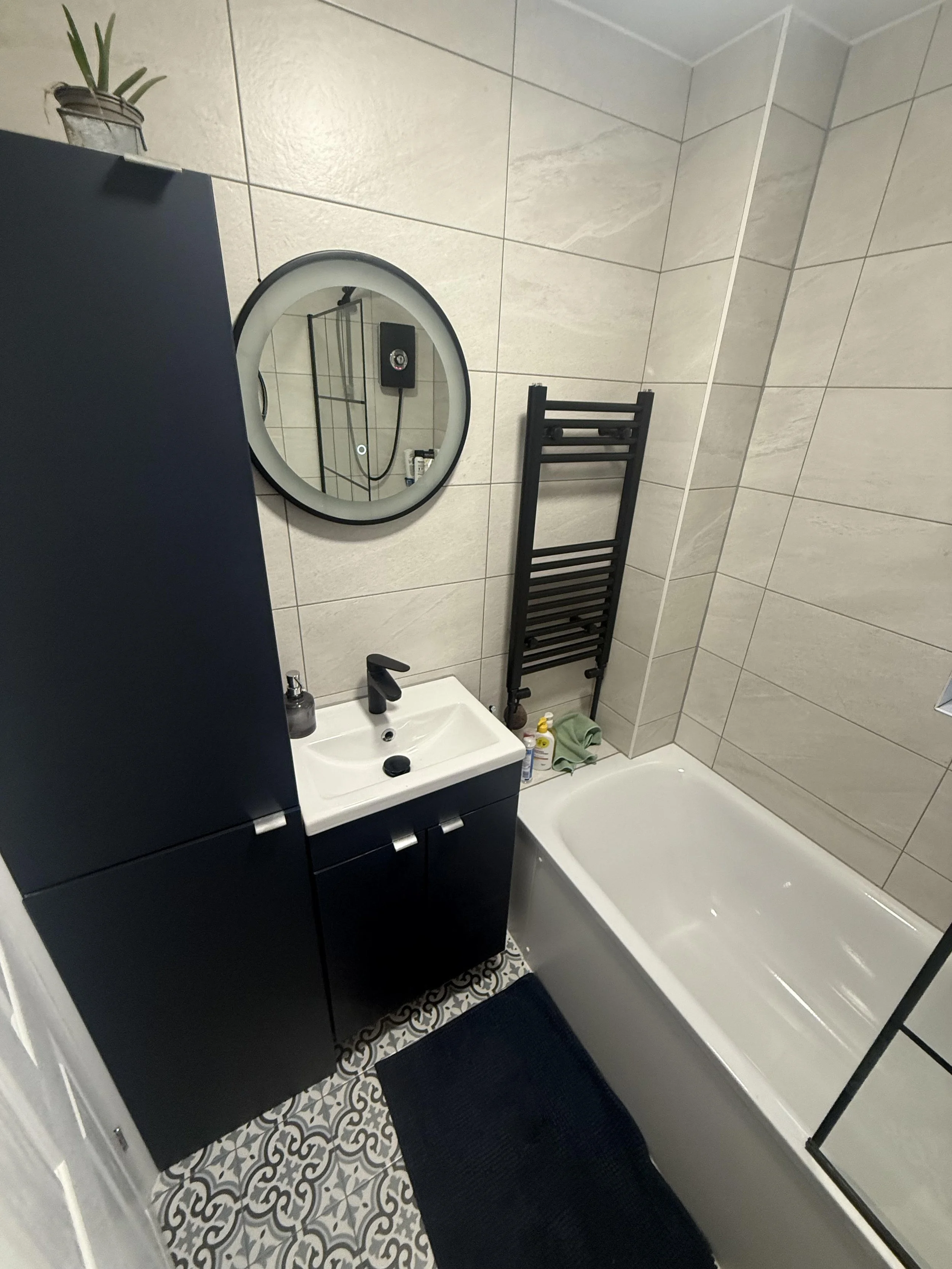 A small modern bathroom with a white bathtub, a black towel radiator, a small white sink with black cabinet, a round mirror, patterned floor tiles, and a black bath mat.