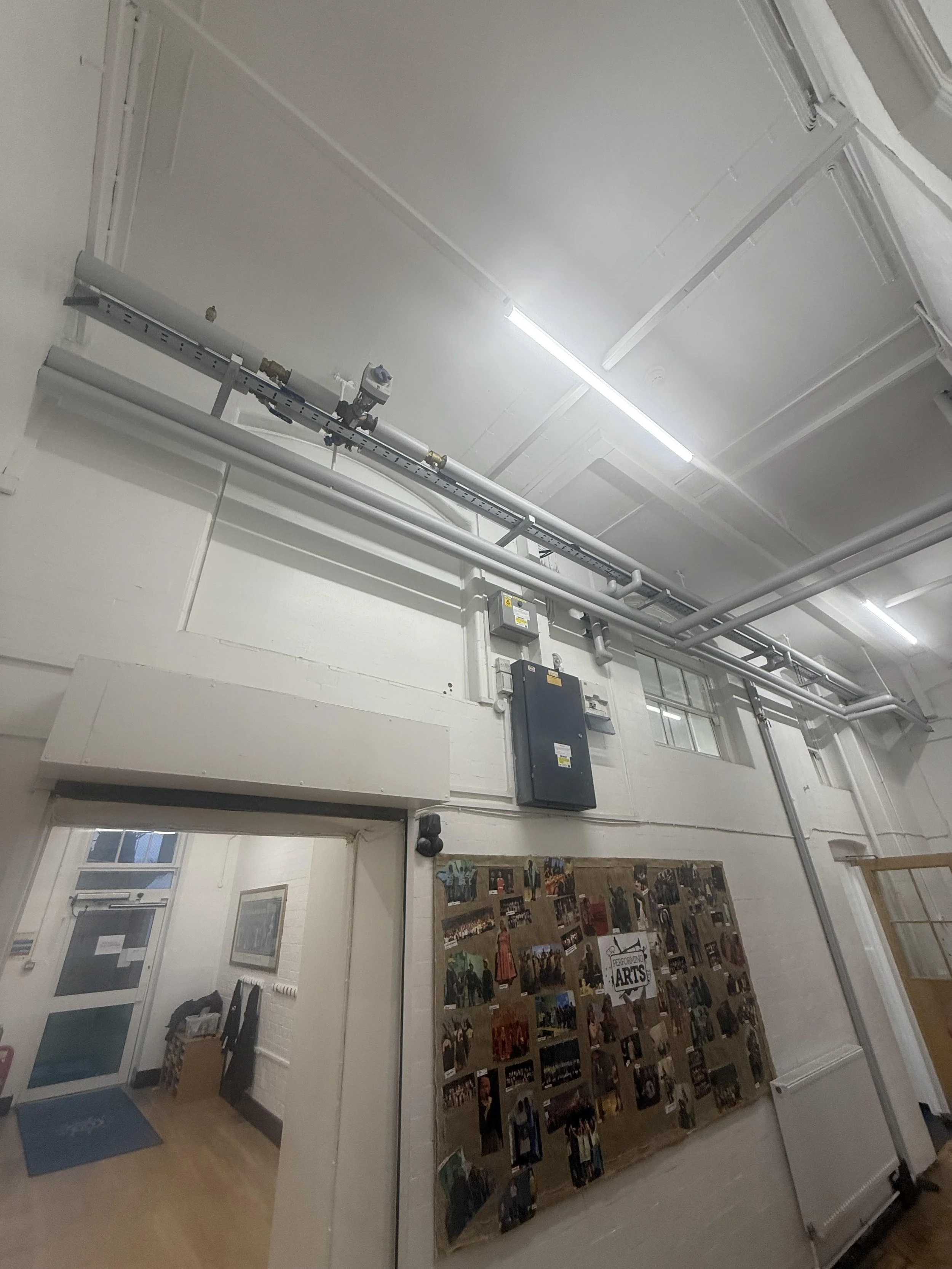 Interior of a room with white walls, ceiling, and exposed pipes and electrical conduit. A black board displays a collage of photos and a sign that reads 'Performing Arts'. A doorway leads to another room with a blue mat and a coat rack.
