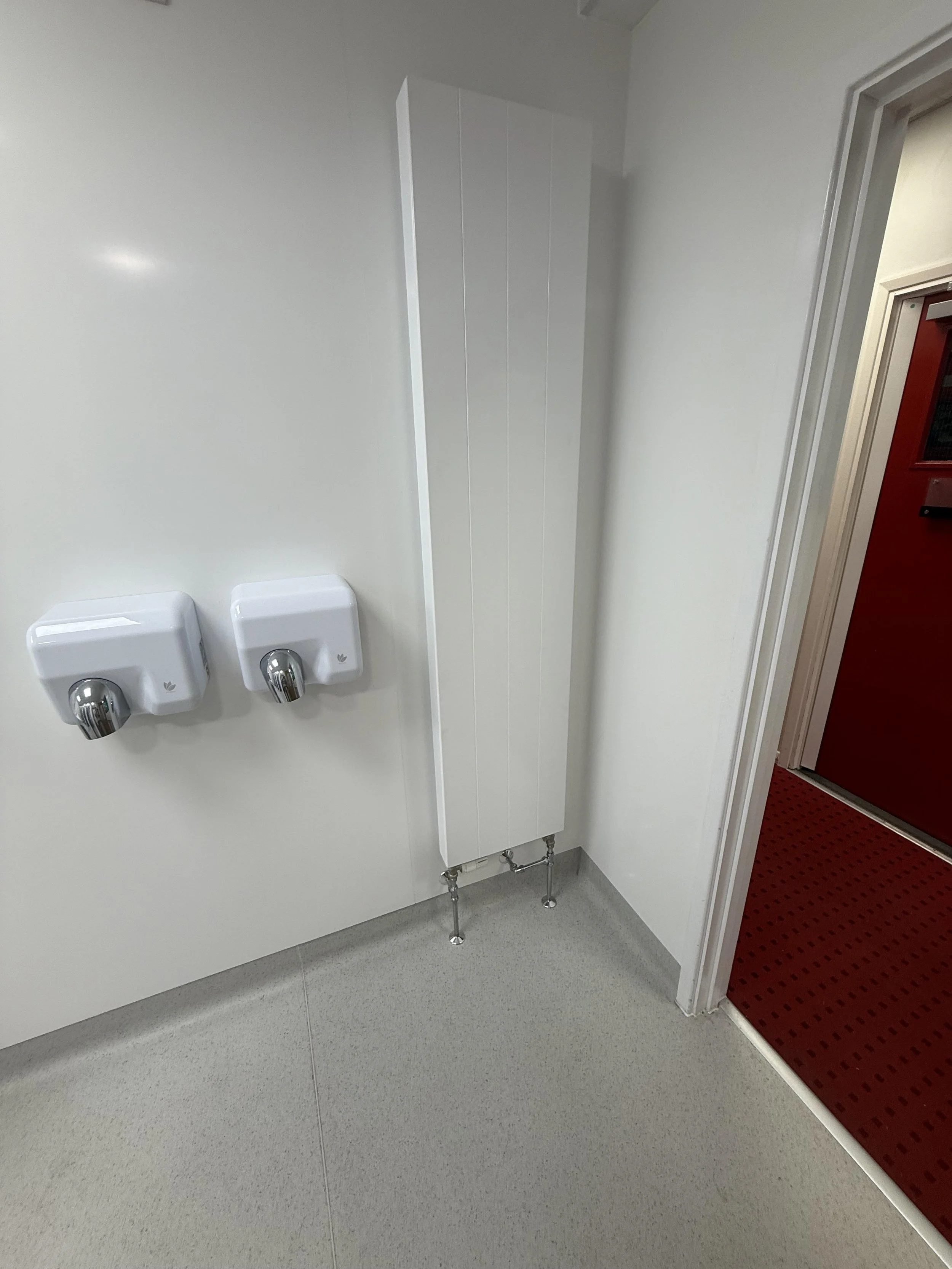 Two soap dispensers on a white wall and a tall, white radiator next to a door with a red mat outside.