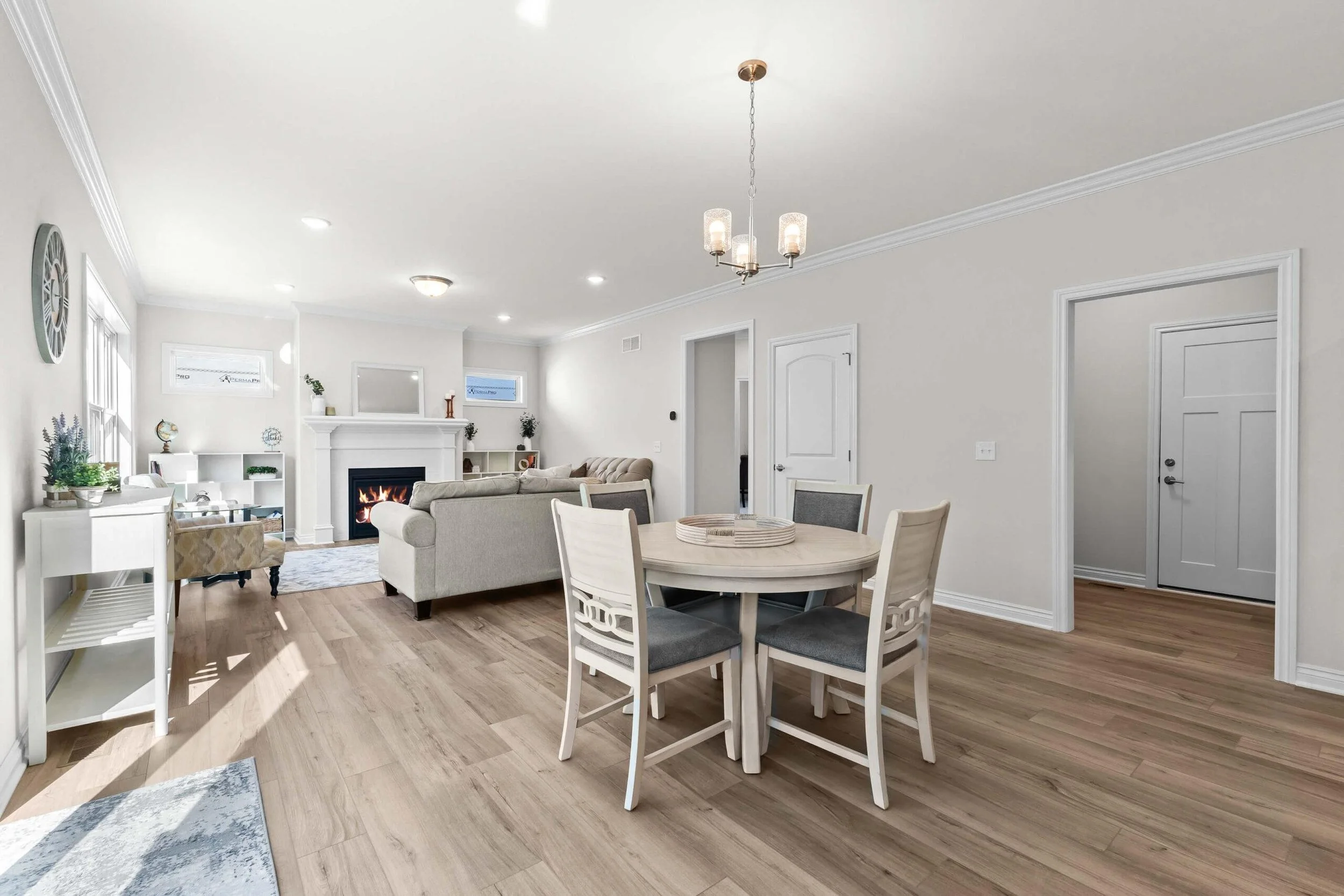 Open-concept living and dining room with a light color palette; white walls, light wood flooring, a round dining table with chairs, a sitting area with a sofa, a fireplace, and multiple windows providing natural light.